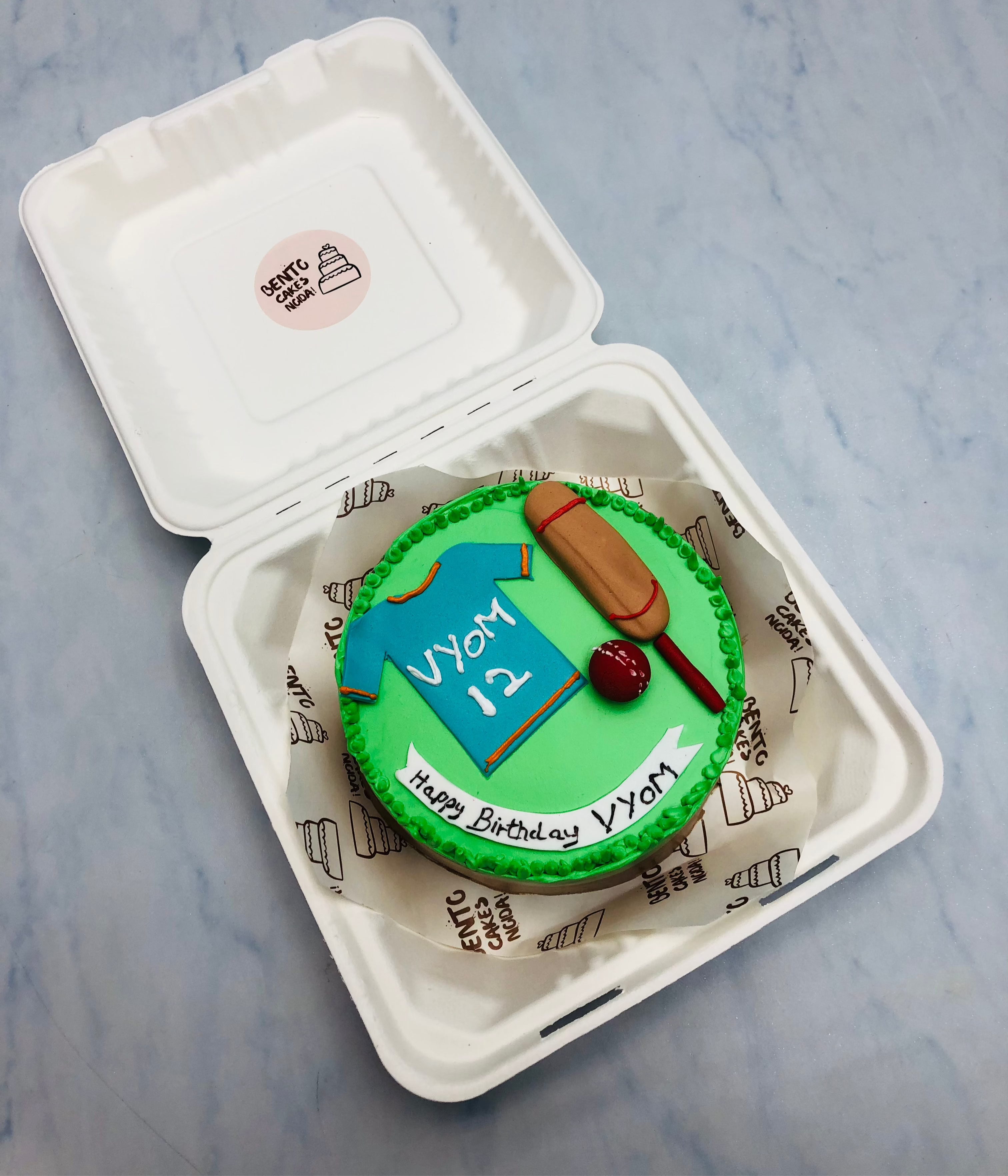 A cricket theme green bento cake with a fondant bat, ball and tshirt.