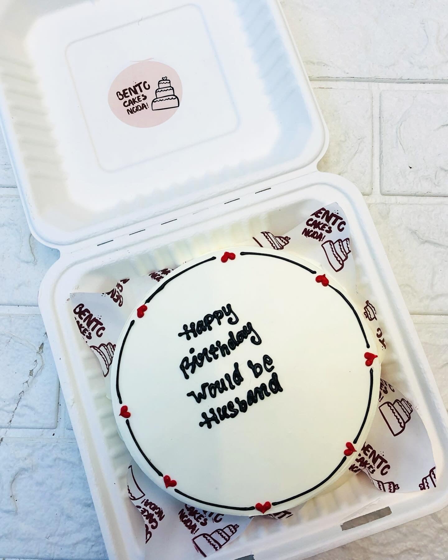 A beautifully crafted white cake with 'Happy Birthday would be Husband' inscribed in black icing, adorned with delicate red heart decorations. The cake is elegantly placed inside a white bento box.
