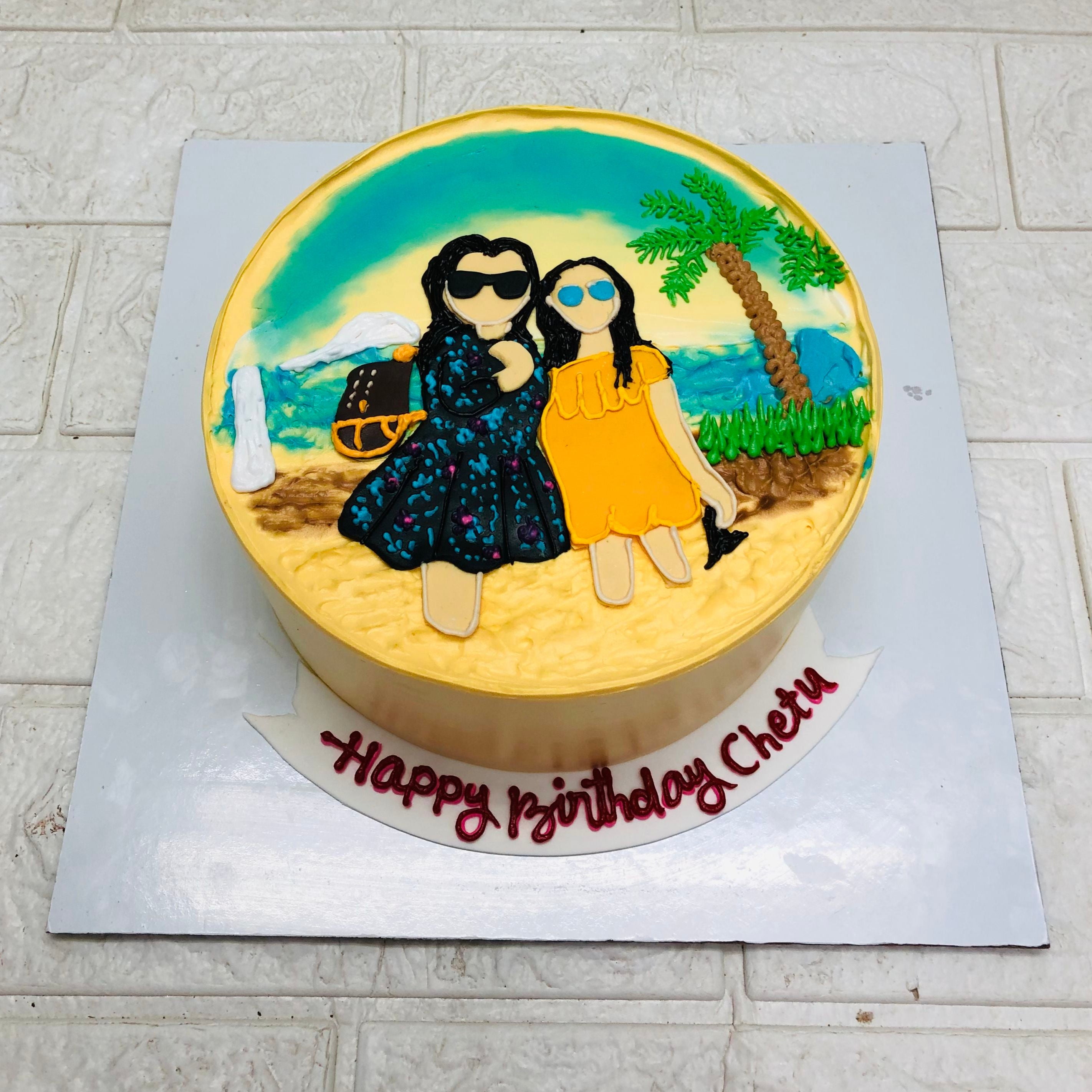 A round birthday cake with a beach-themed design, depicting two figures standing on a beach with a palm tree and ocean waves in the background. The cake has "Happy Birthday Chetu" written in red icing on a white board at the base showing top view.