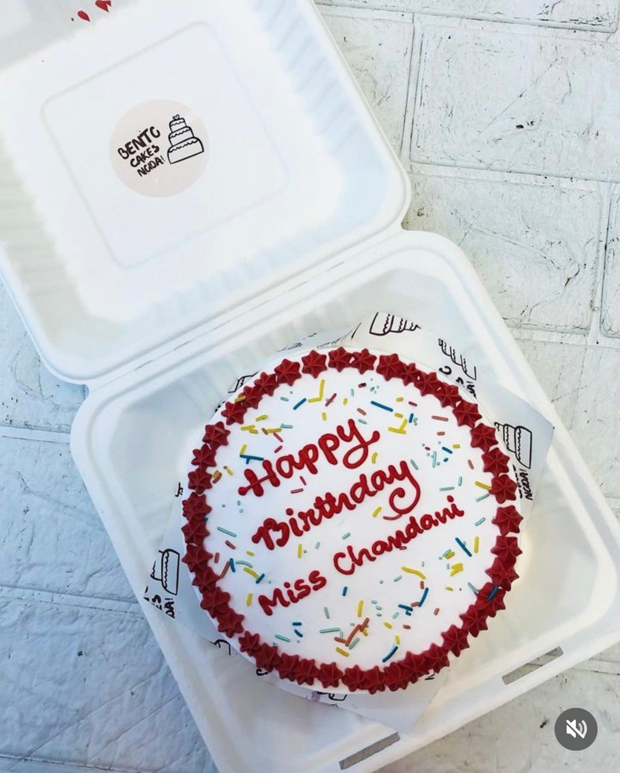 A charming white birthday cake with red icing borders and colorful sprinkles, placed inside a stylish white bento cake box. The cake features the message 'Happy Birthday Miss Chandani' in red icing, adding a personalized touch.
