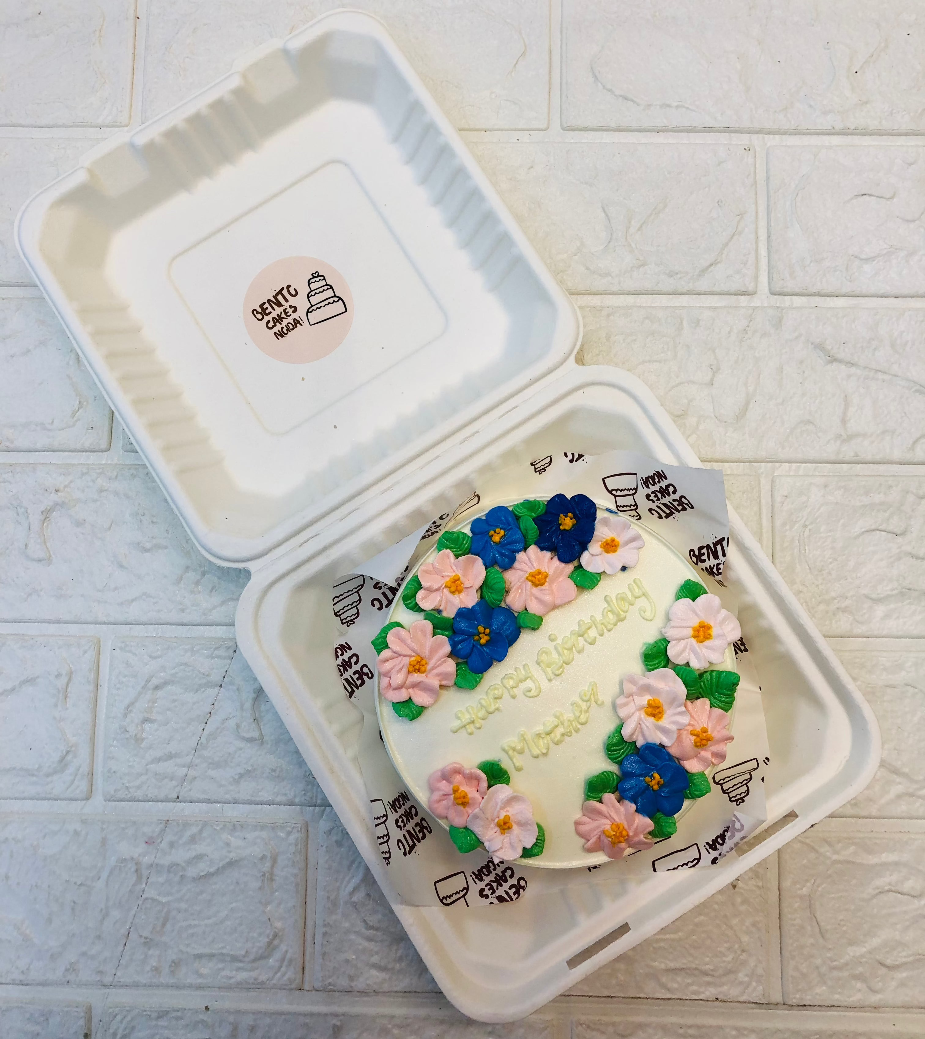 - A beautifully crafted small round cake inside a white bento box, adorned with pink and blue flowers and green leaves. The cake features a personalized message, "Happy Birthday Mother" written in yellow icing.