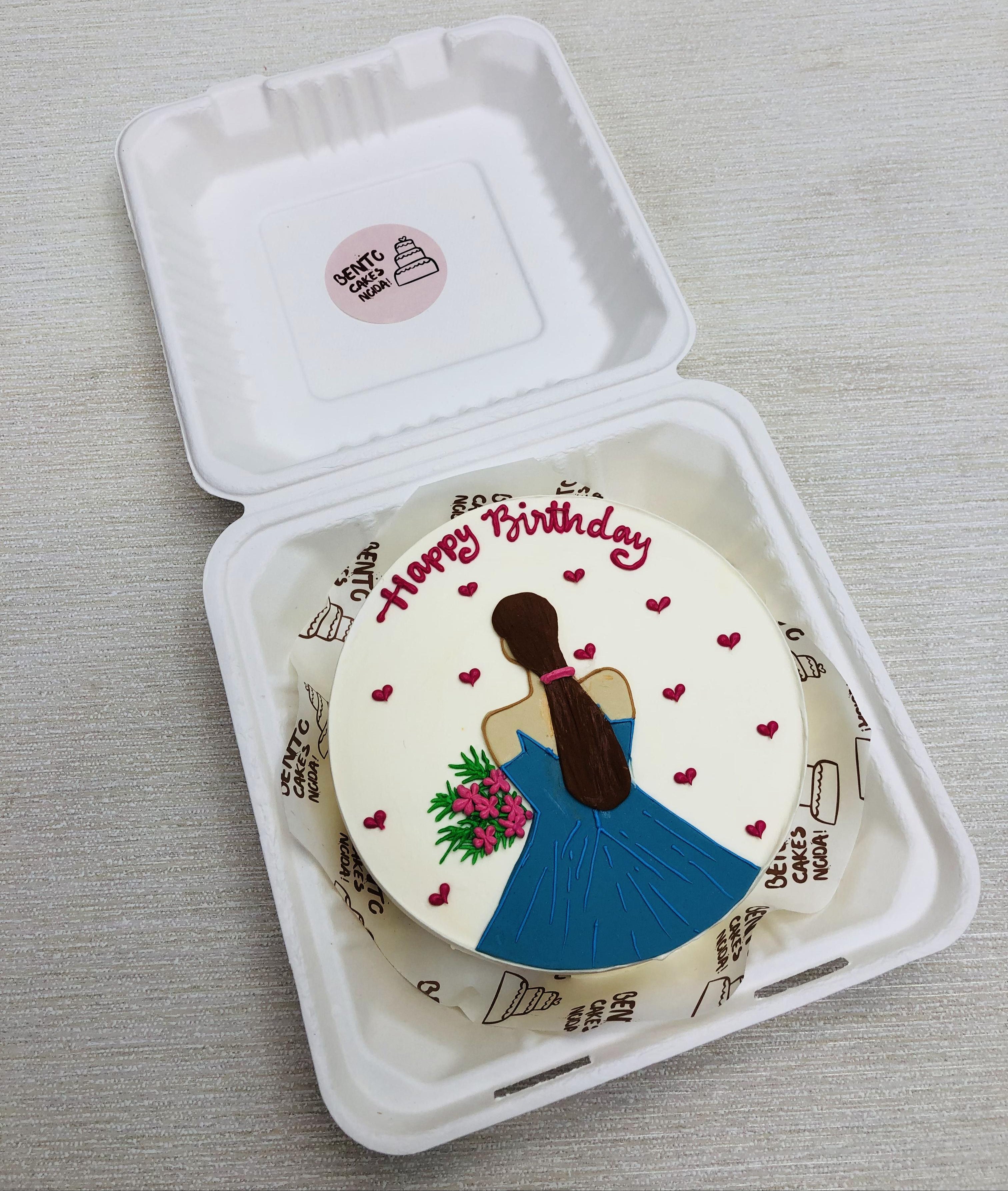 A beautifully decorated birthday cake featuring an illustration of a girl with long brown hair wearing a blue dress and holding a bouquet of flowers. The cake is placed inside a white box with a label that reads "SWEET CAKE SHOP." The cake has "Happy Birthday" written in red icing at the top, surrounded by small red hearts.