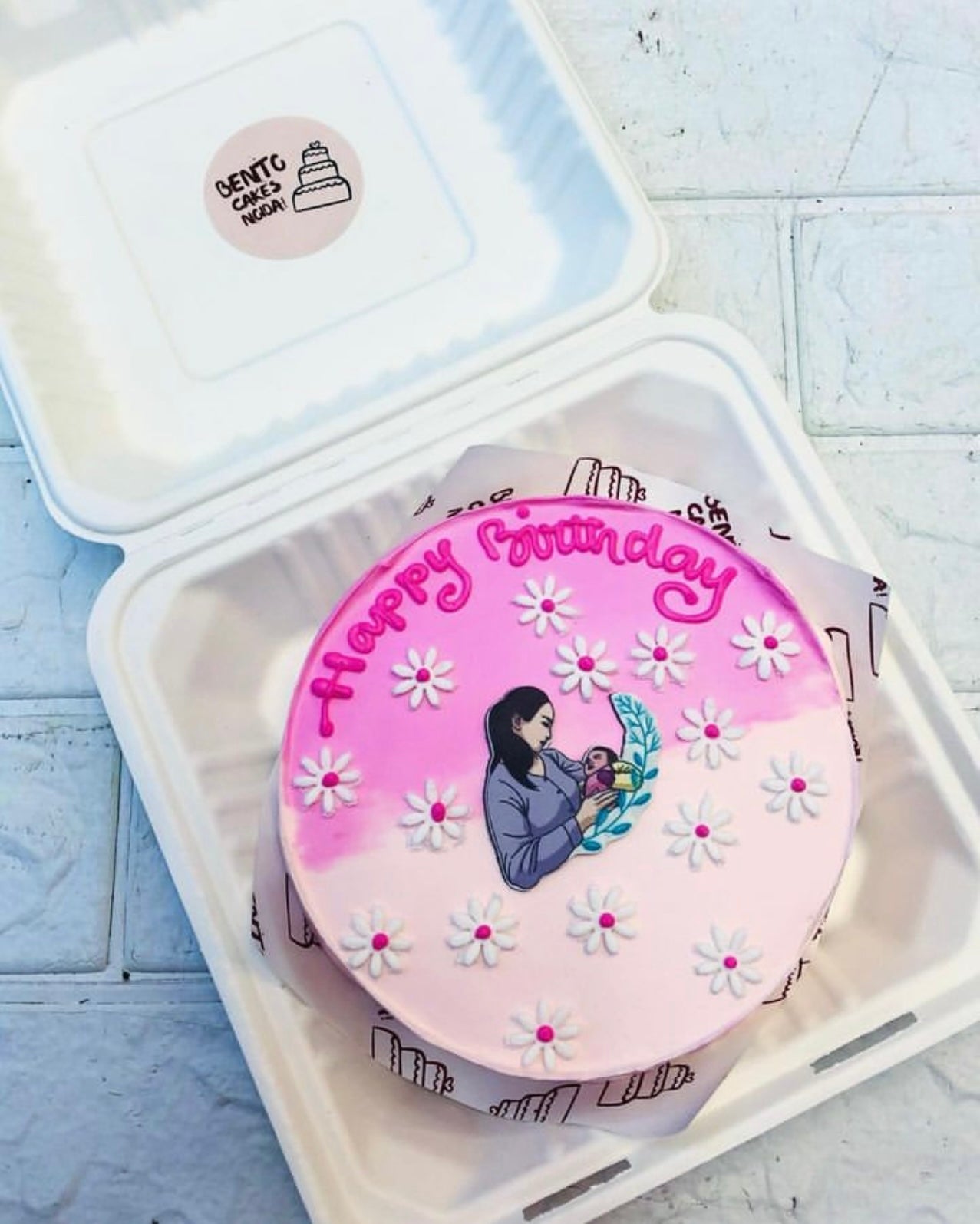 A beautifully decorated bento cake inside a white box. The cake has a pink and white gradient background with the text "Happy Birthday" written in pink icing at the top. The cake features an illustration of a person holding a baby, surrounded by white daisy flowers with pink centers.
This title captures the essence of the cake's personalized artwork and elegant decoration. Let me know if you'd like any tweaks!
