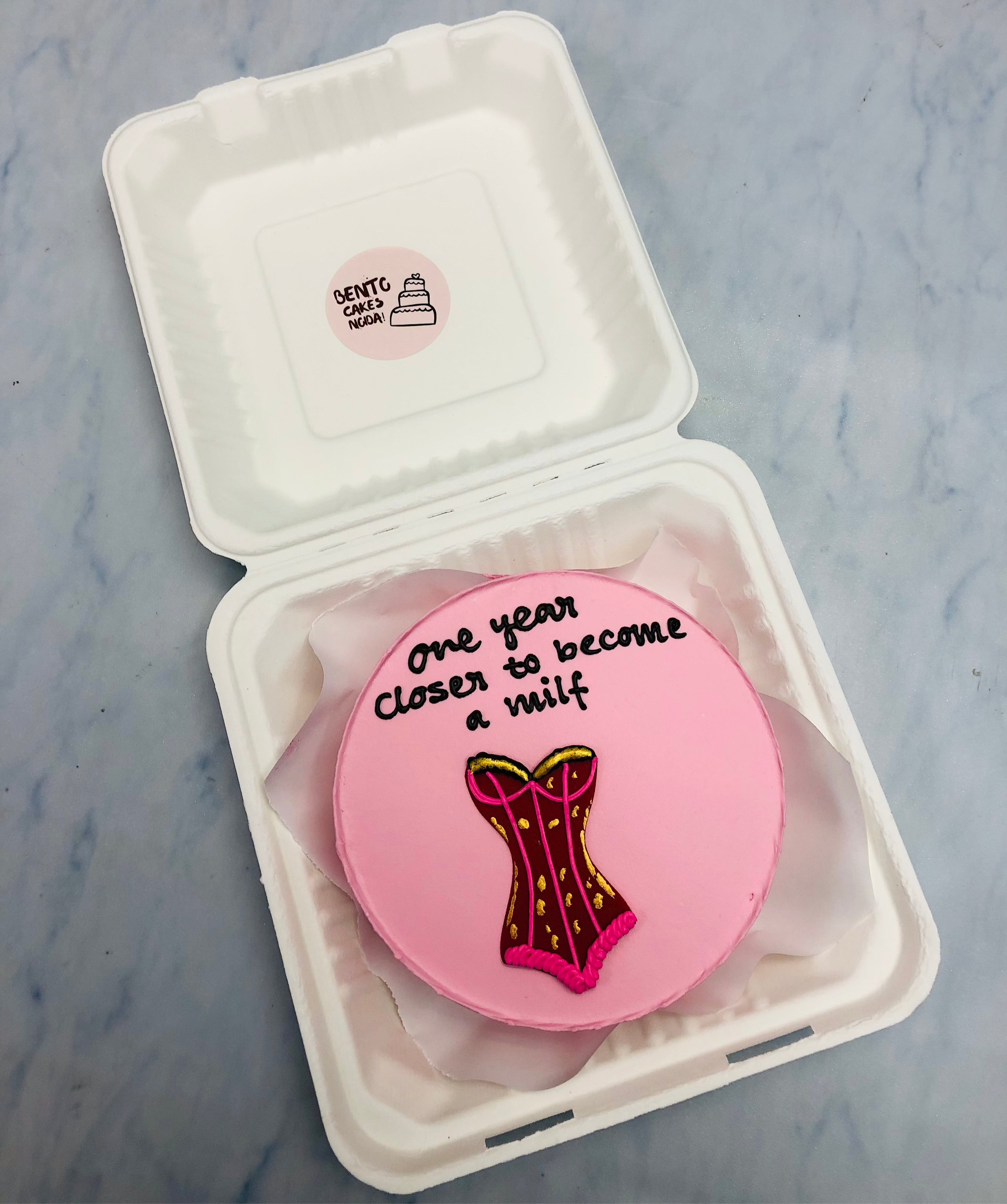 A unique pink birthday cake featuring a corset design and a humorous message, “One year closer to becoming a MILF,” placed inside a bento box.