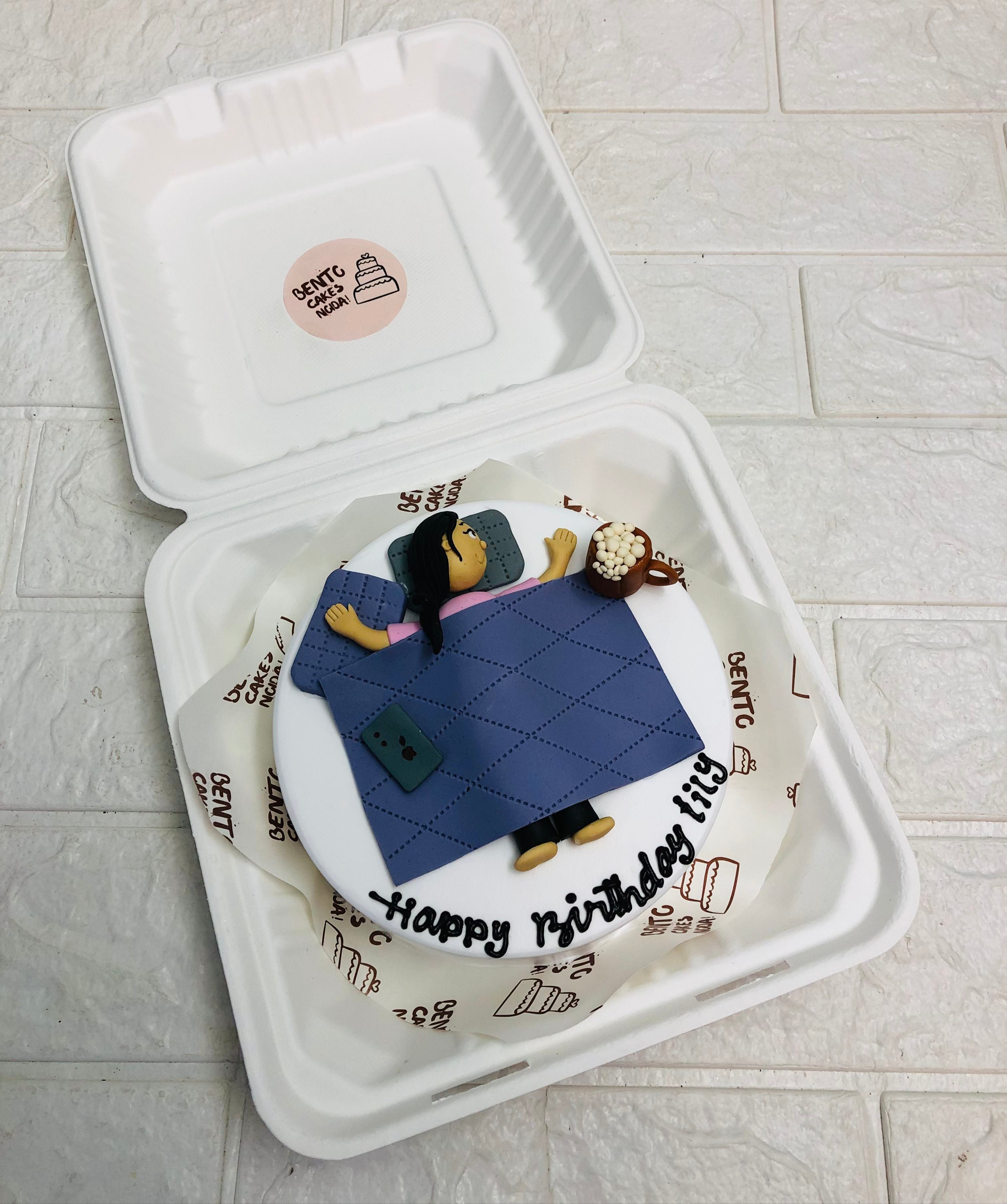 A bento cake in a bento box with a cozy bedtime-themed decoration. The cake features a figure lying in bed with a blue quilt, holding a pillow and a phone, with a cup of popcorn on a bedside table. The words "Happy Birthday Lily" are written on the cake.
