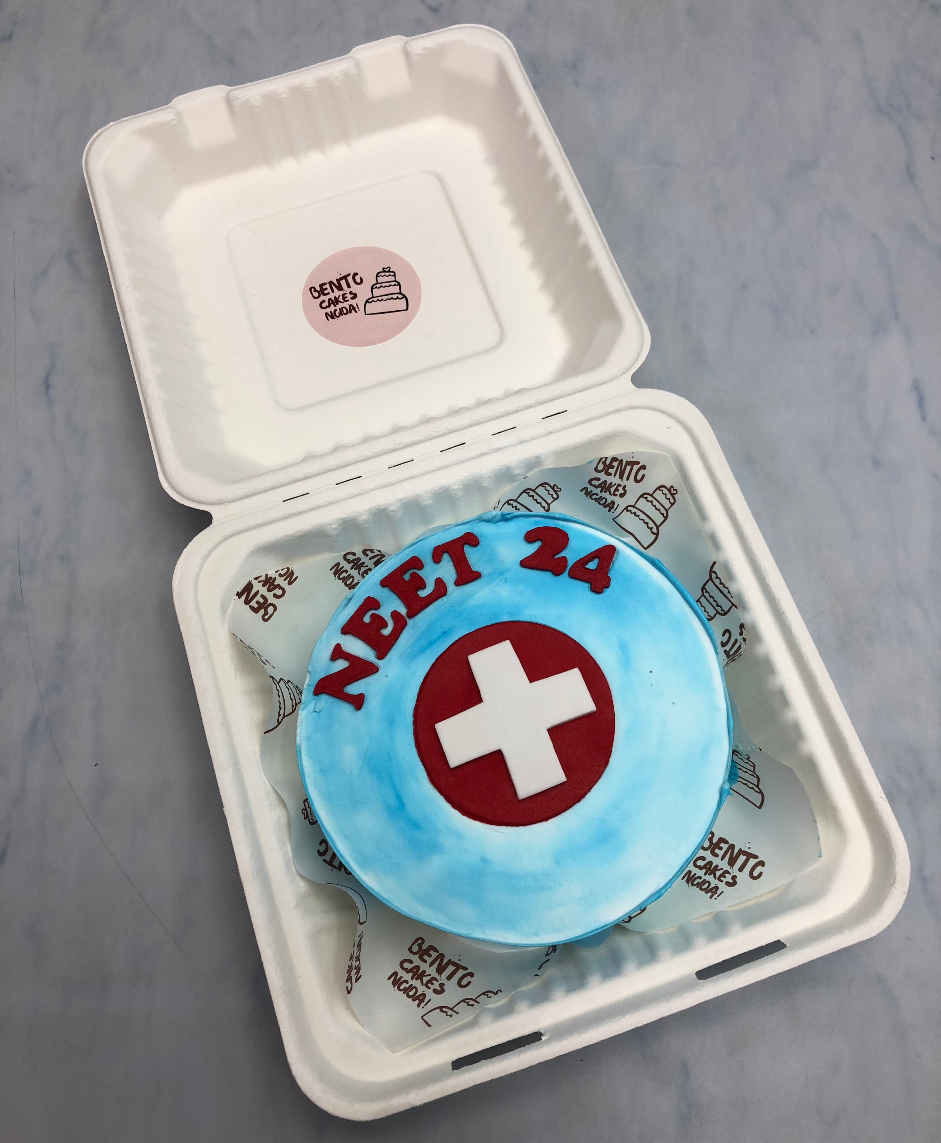 An aesthetic blue bento cake is designed for being a doctor and having a text "NEET 24" with red fondant alphabets.