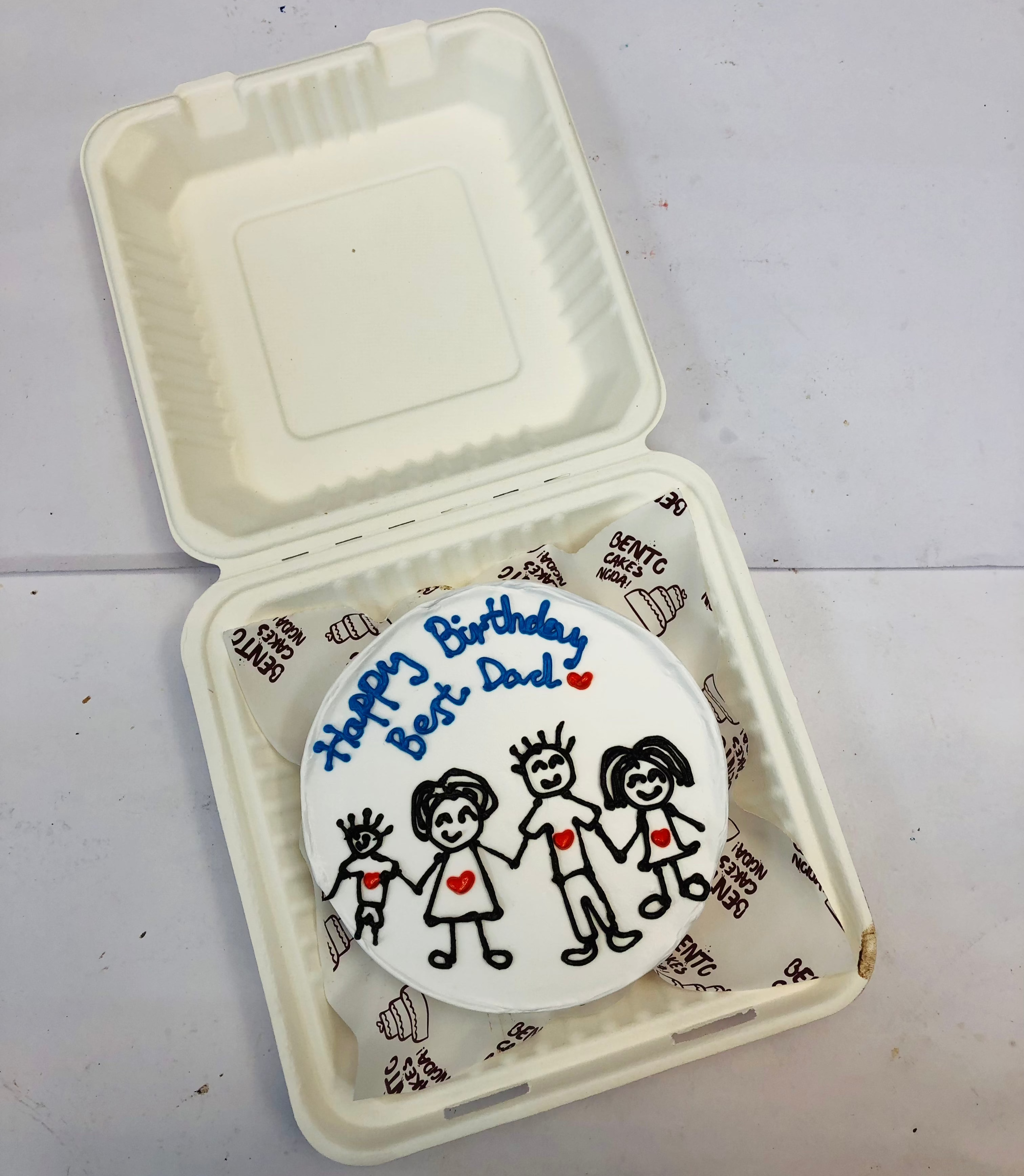A birthday cake inside a white box, decorated with a hand-drawn family of four holding hands. The message 'Happy Birthday Best Dad' is written in blue icing, with red hearts on each family member’s chest.