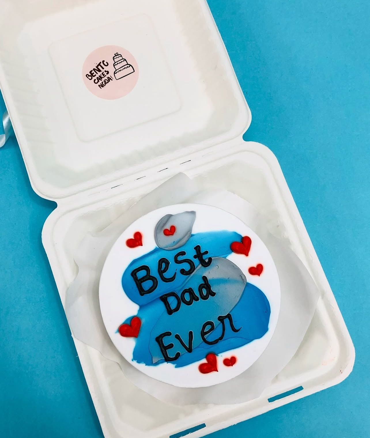 A white cake with blue and gray decorative elements, featuring “Best Dad Ever” in black icing, surrounded by red heart accents.