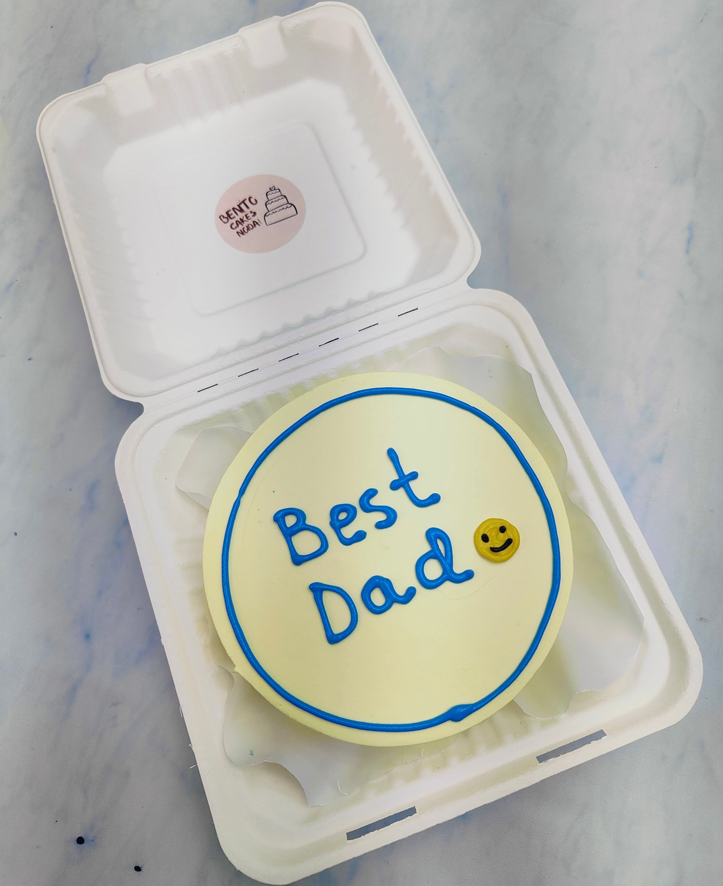 A white bento cake having a blue line border with a blue text "Best Dad" with a smiley emoji on cake.