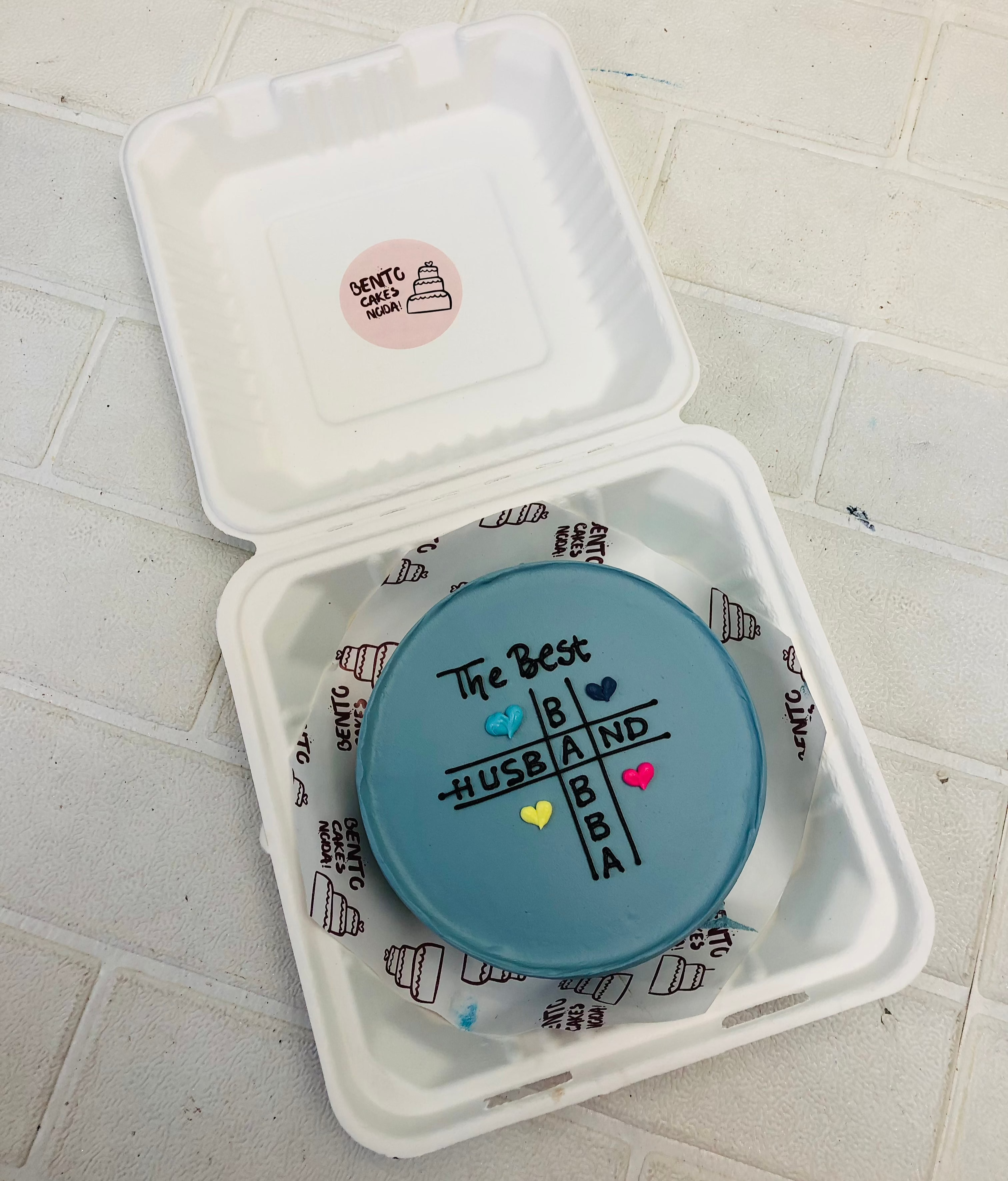 A dark grey bento cake is designed in a tic tac tow design for the best "HUSBAND" "BABBA".