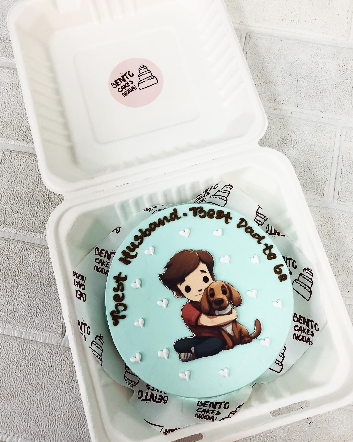 A light blue bento cake aving a boy/man with his dog printed on cake and decoated with white hearts.