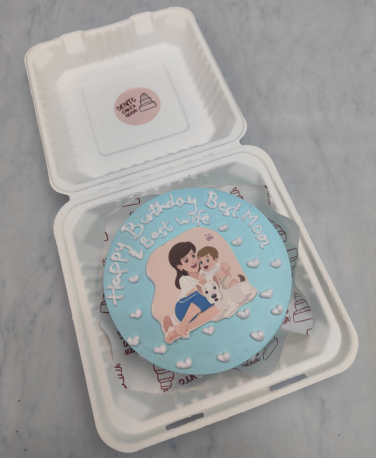 A light blue bento cake with a mom and baby print on it with her pet puppy. The cake shows best mom and wife emotion.