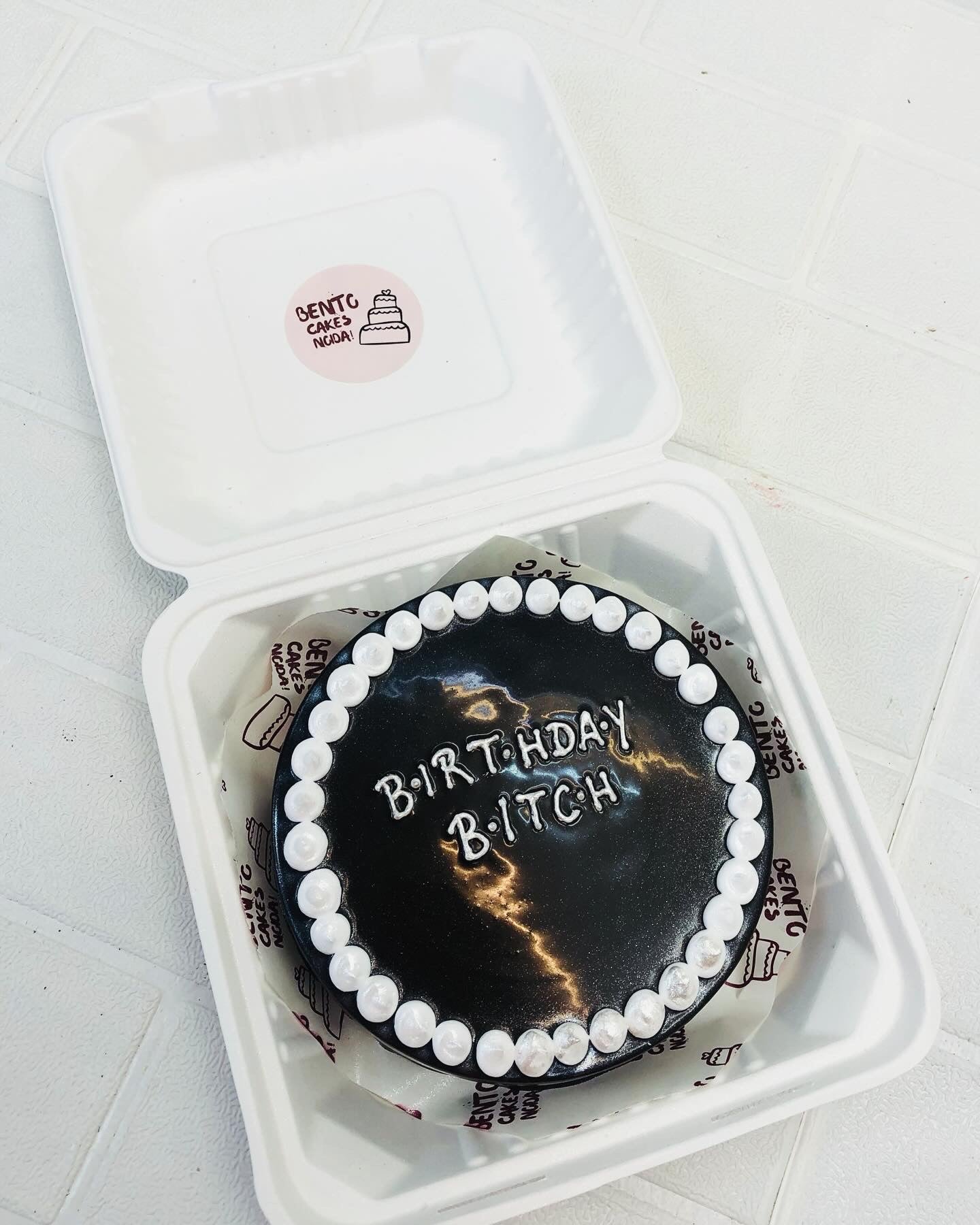 A black icing on a bento cake with white dotted boundary and a text "BIRTHDAY BITCH" written with white icing.