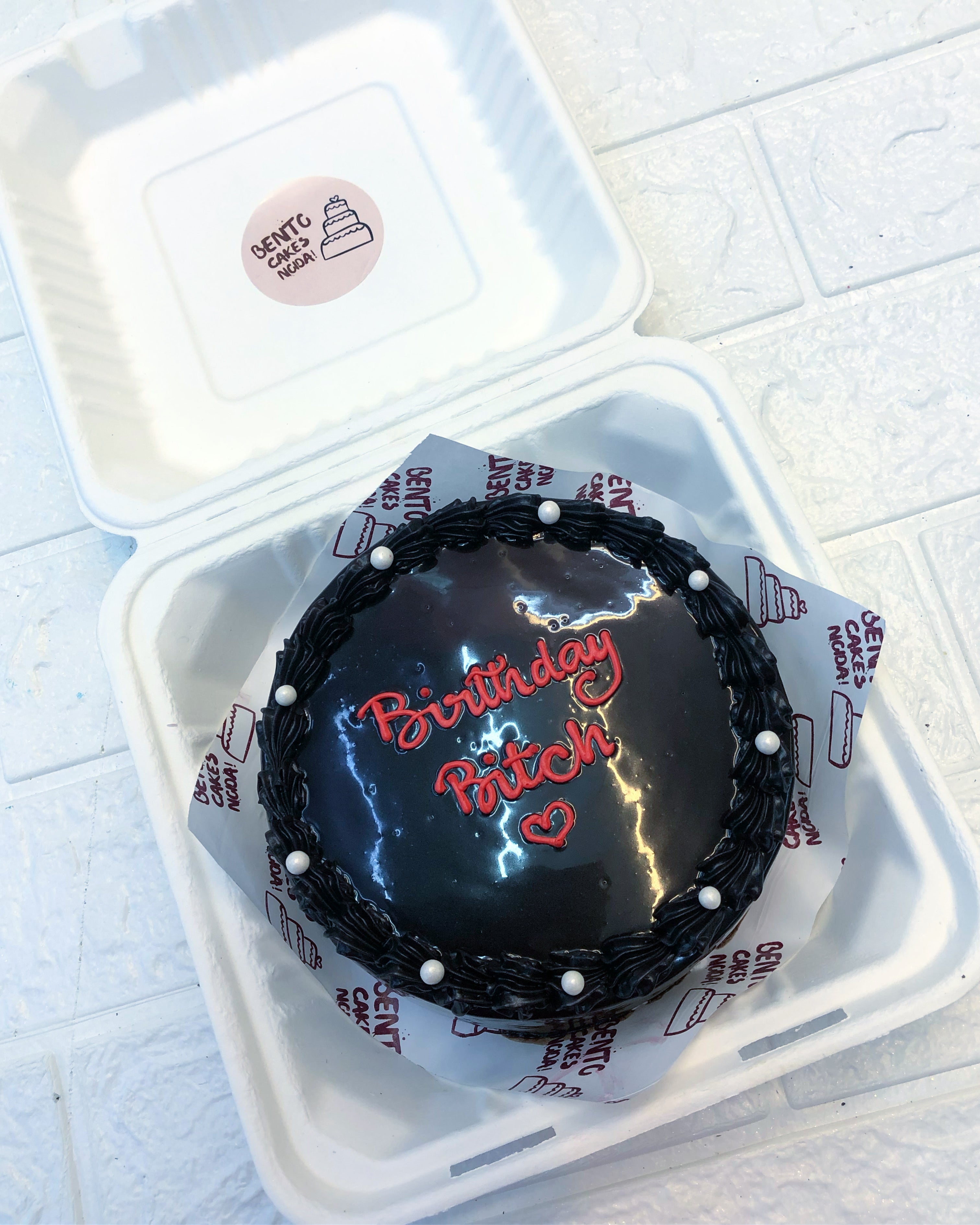 A round birthday cake with a glossy black icing, decorated with white pearls around the edge and red lettering that reads 'Birthday Bitch' with a heart underneath.