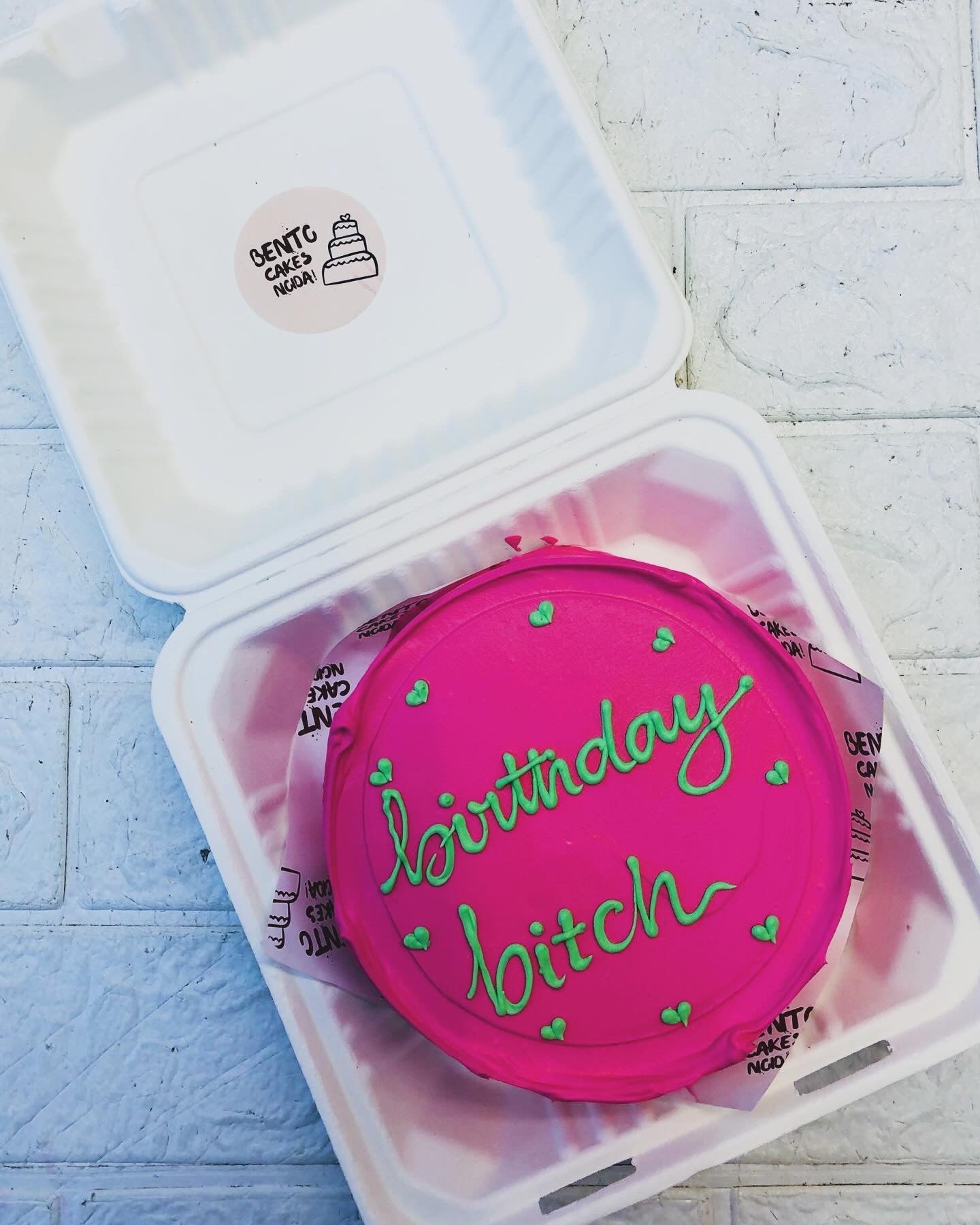 A pink birthday cake with green piping and playful lettering that says "birthday bitch," adorned with small green hearts. The cake sits inside an open white bento cake box on a tiled surface.