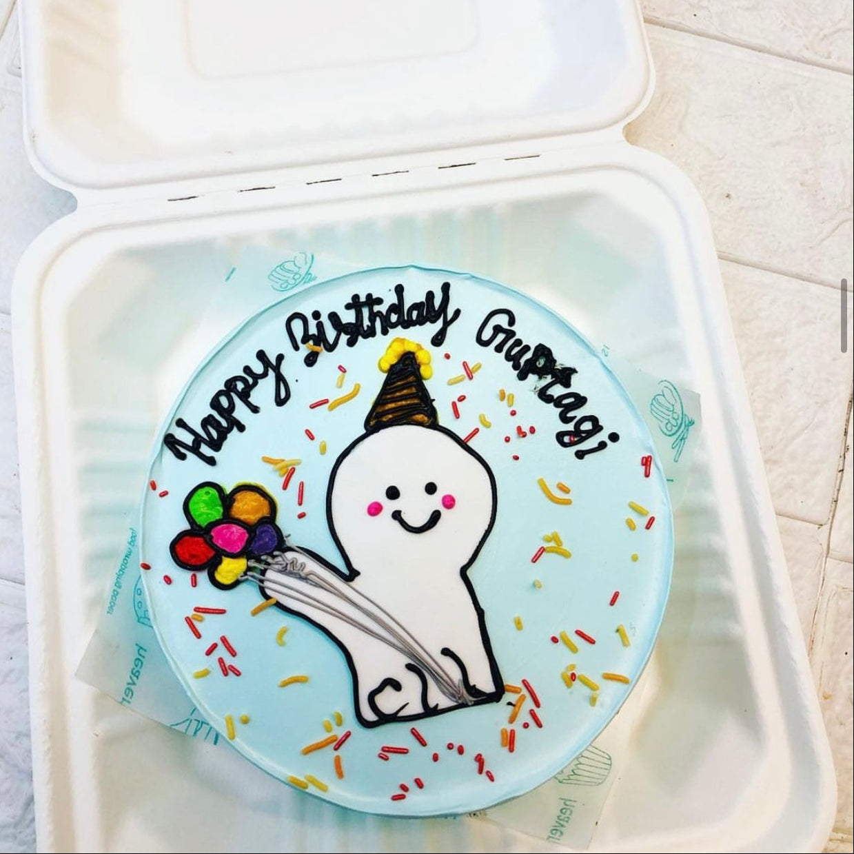 A light blue birthday cake featuring a cute cartoon character holding colorful balloons and wearing a party hat. The cake is decorated with colorful sprinkles and the text "Happy Birthday Guptagi" written in black icing. 