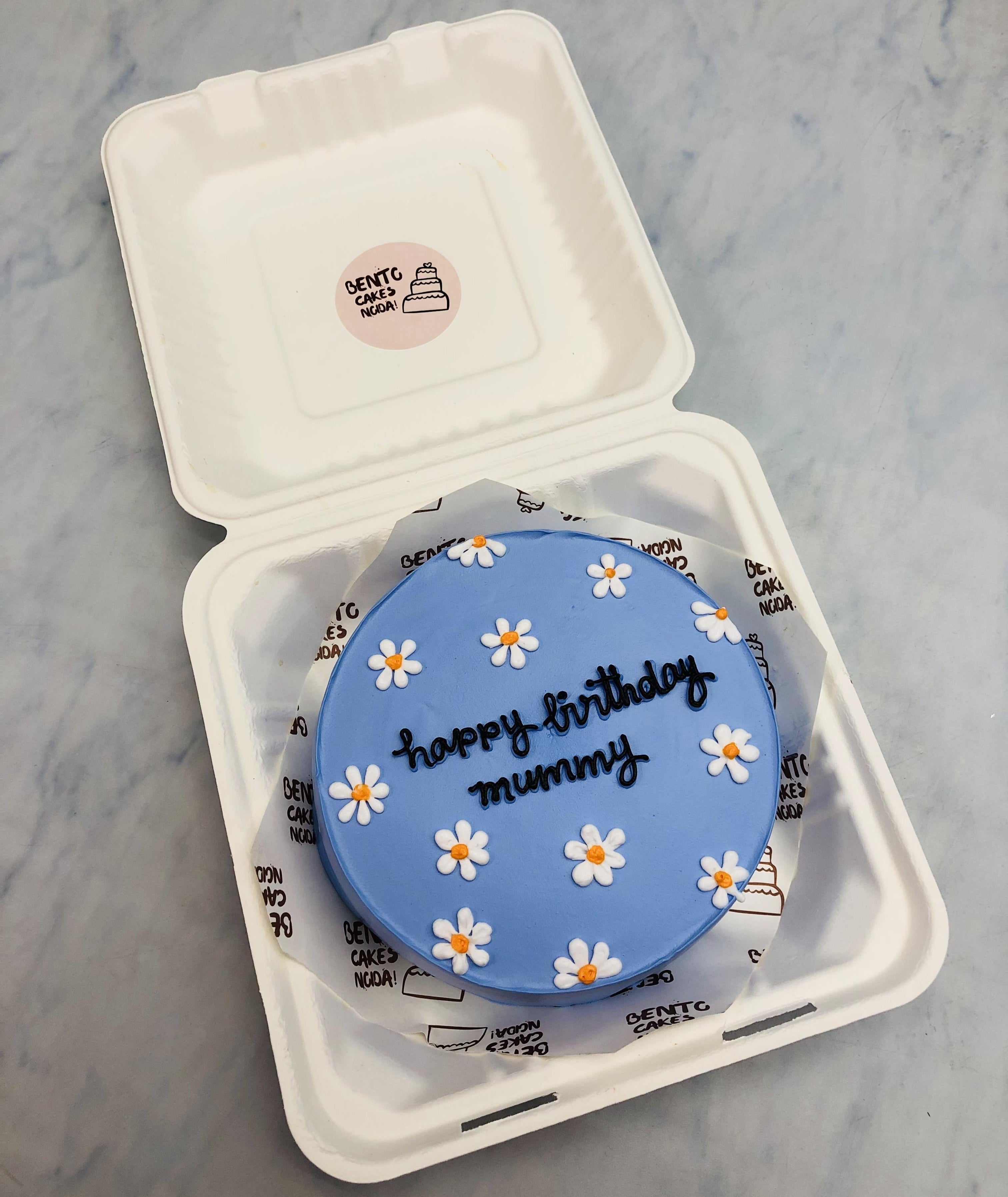 A charming blue round cake adorned with delicate white and yellow daisies, presented inside a white bento cake box. The cake features 'happy birthday mummy' written in black icing, making it a heartfelt and visually stunning birthday treat."
