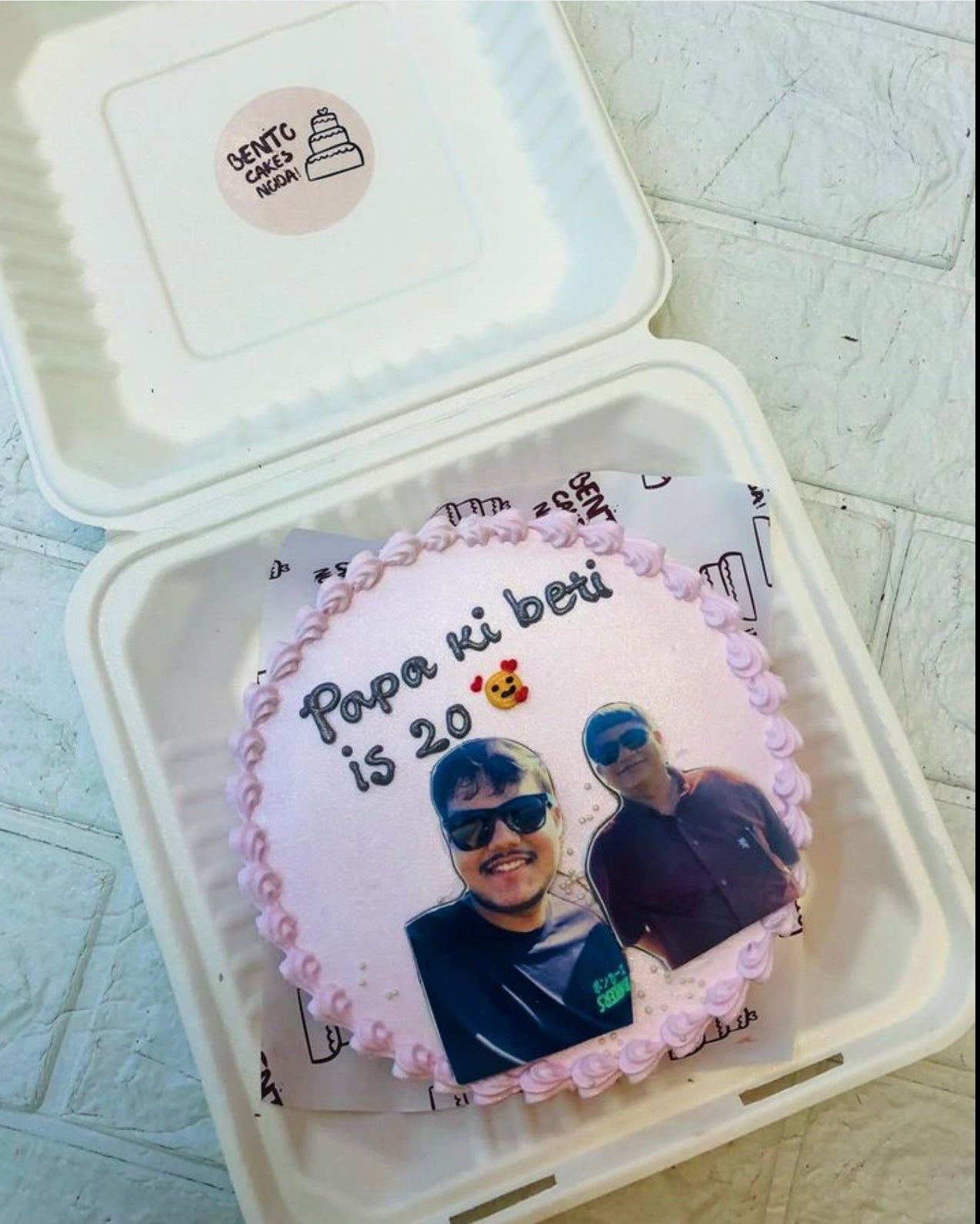 A round birthday cake with light pink frosting, decorated with a photo of two people and the text 'Papa ki beti is 20' along with a small bear emoji.
