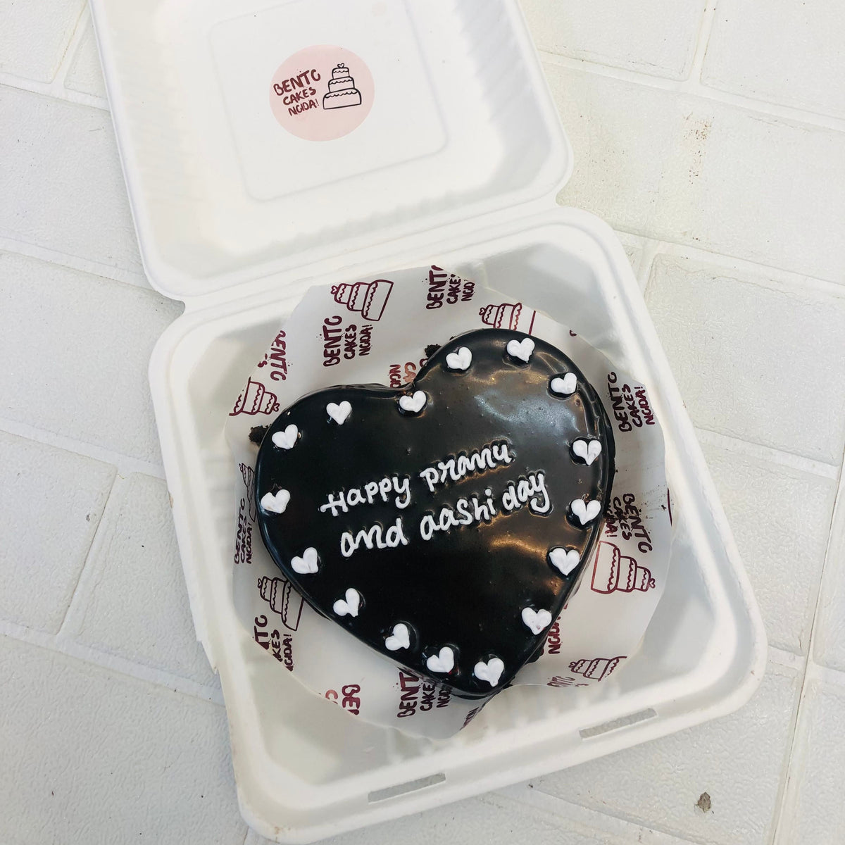 A black icing cake with white haerts on its surrounding and a text with white color on it.
