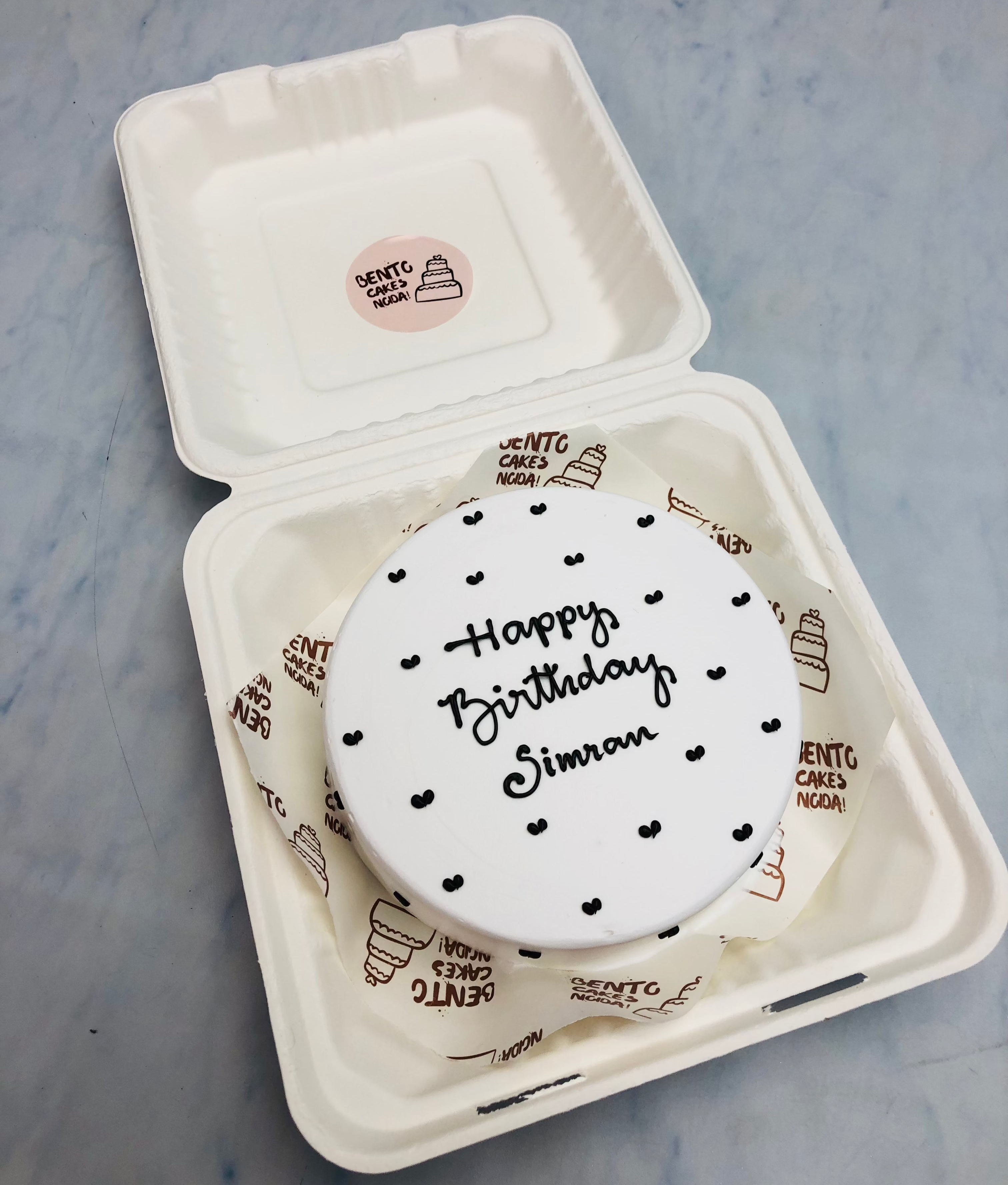 A white round bento cake having little black hearts design all over the cake and a text "Happy Birthday Simra" written with black icing.
