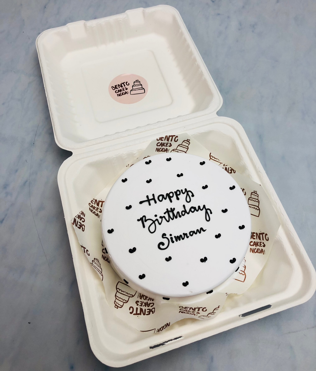 A white round bento cake having little black hearts design all over the cake and a text "Happy Birthday Simra" written with black icing.
