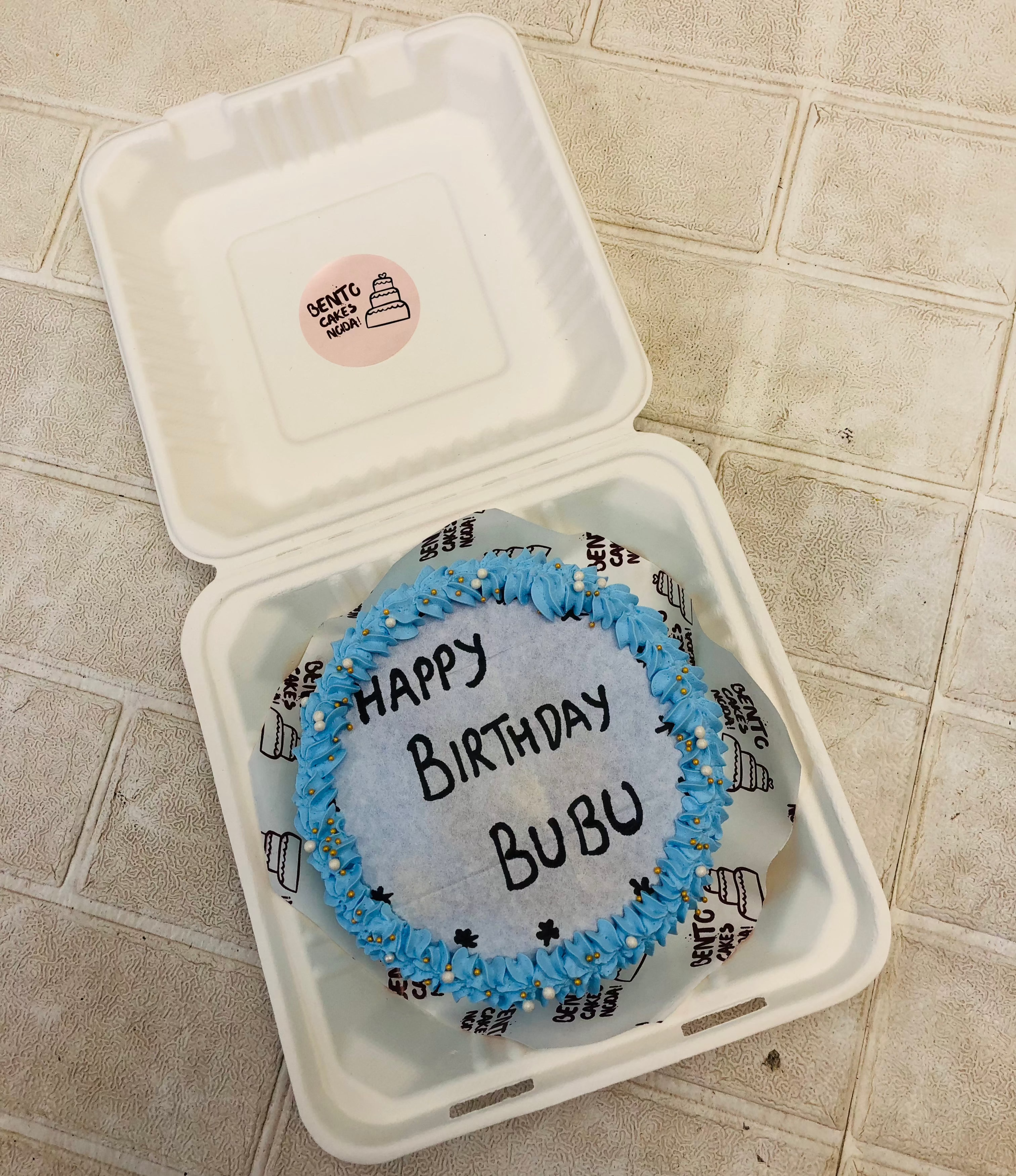 A blue floral design fire bento cake having a text "HAPPT BIRTHDAY BUBU" written on fire paper.