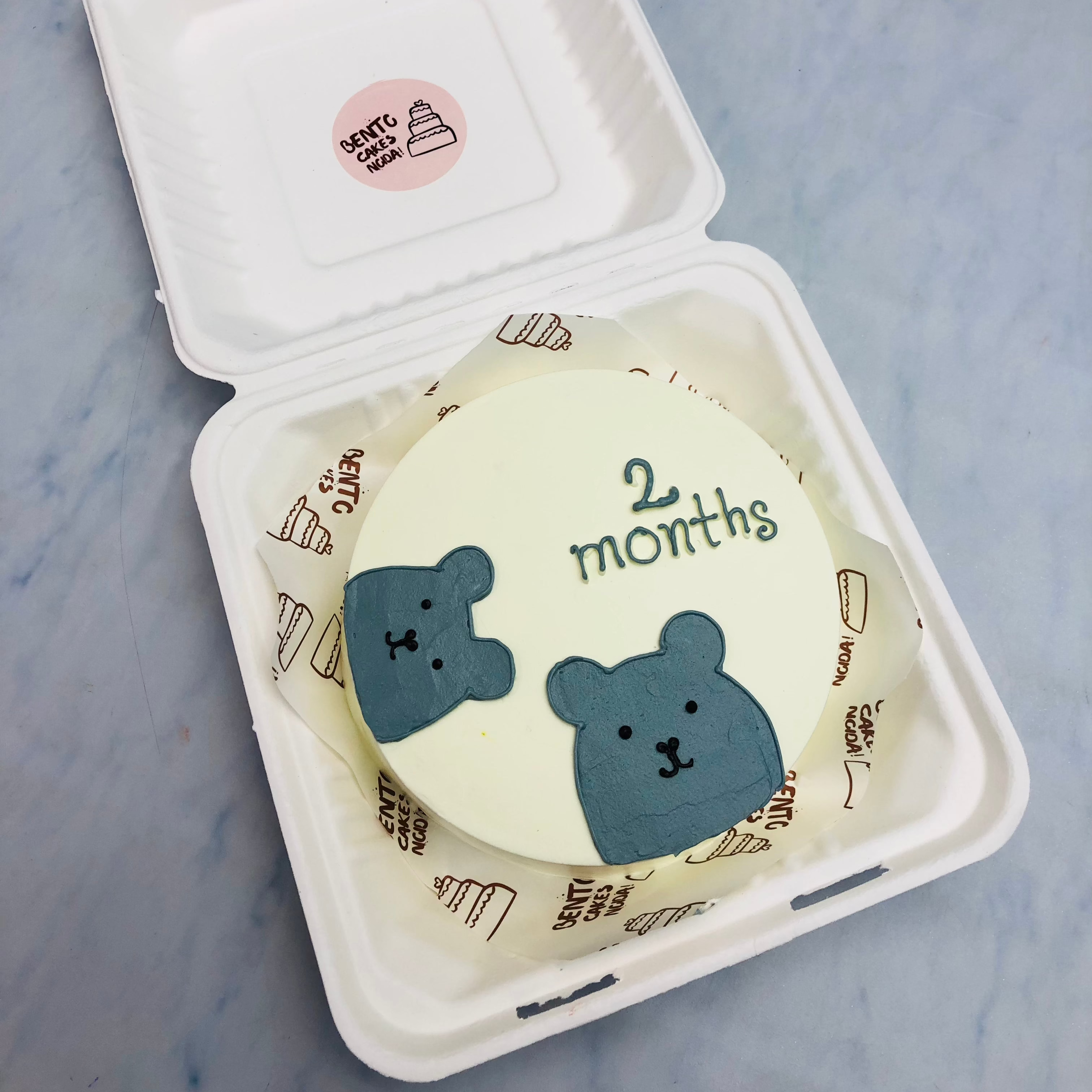 An offwhite bento cake with 2 grey bears and a text "2 months" written on  it.