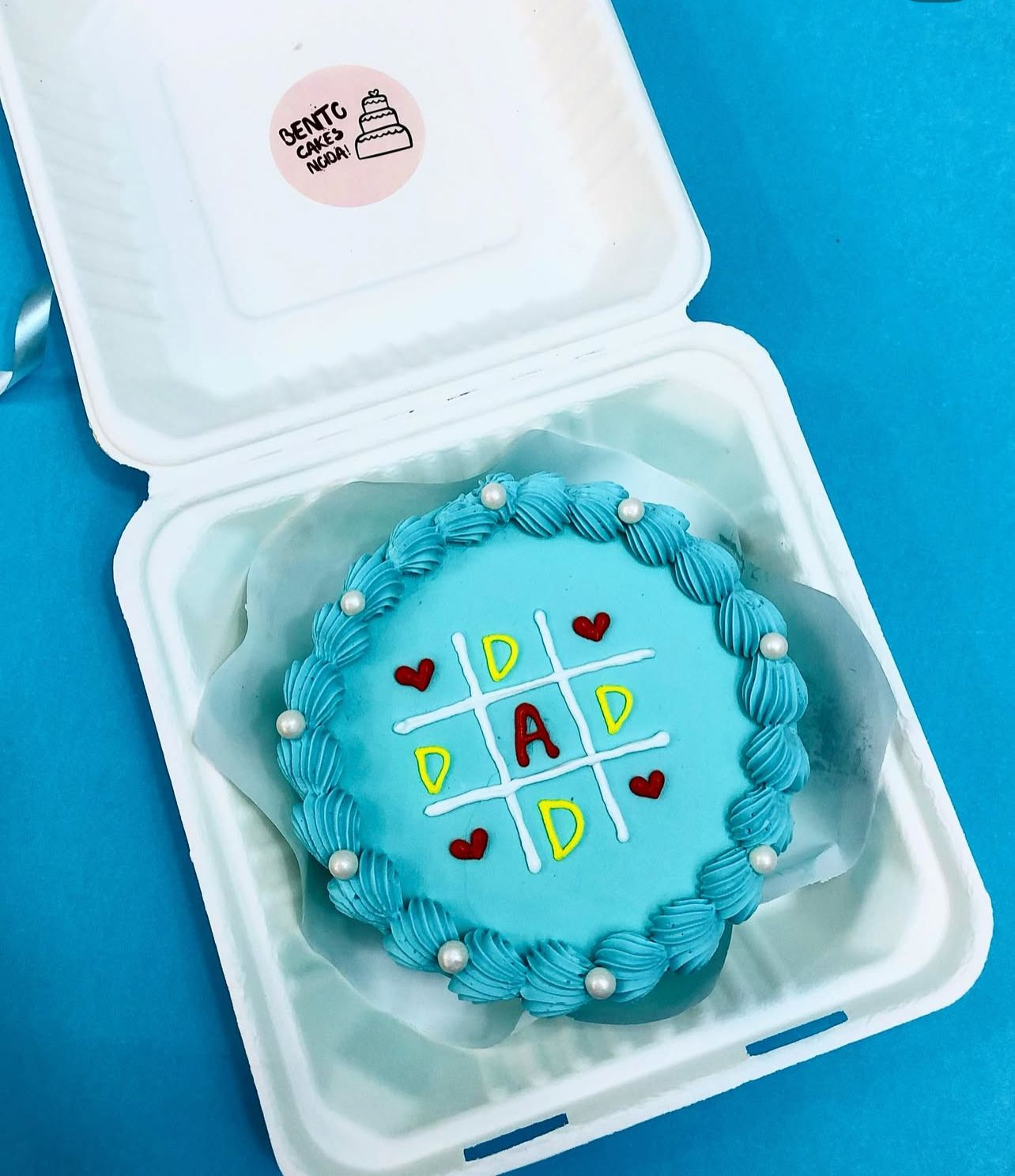 A blue cake featuring a tic-tac-toe design with the word "DAD" in yellow icing and the letter "A" in red icing at the center. The grid is outlined in white icing, and small red hearts surround it. The cake is decorated with blue frosting swirls and pearl-like accents.