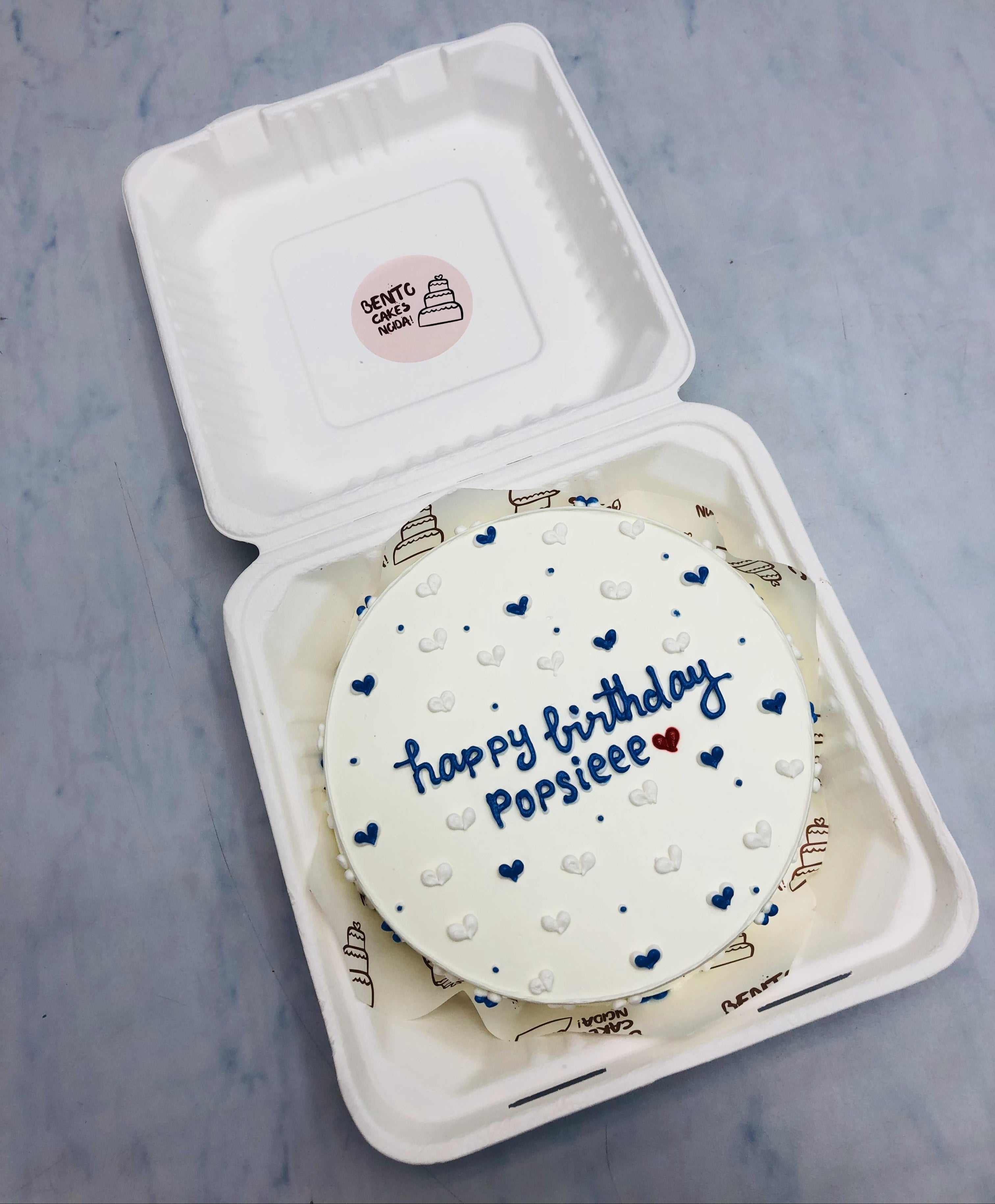 A round birthday cake with white frosting, decorated with blue and white hearts, and the message 'happy birthday Popsieee' written in blue icing with a small red heart. 