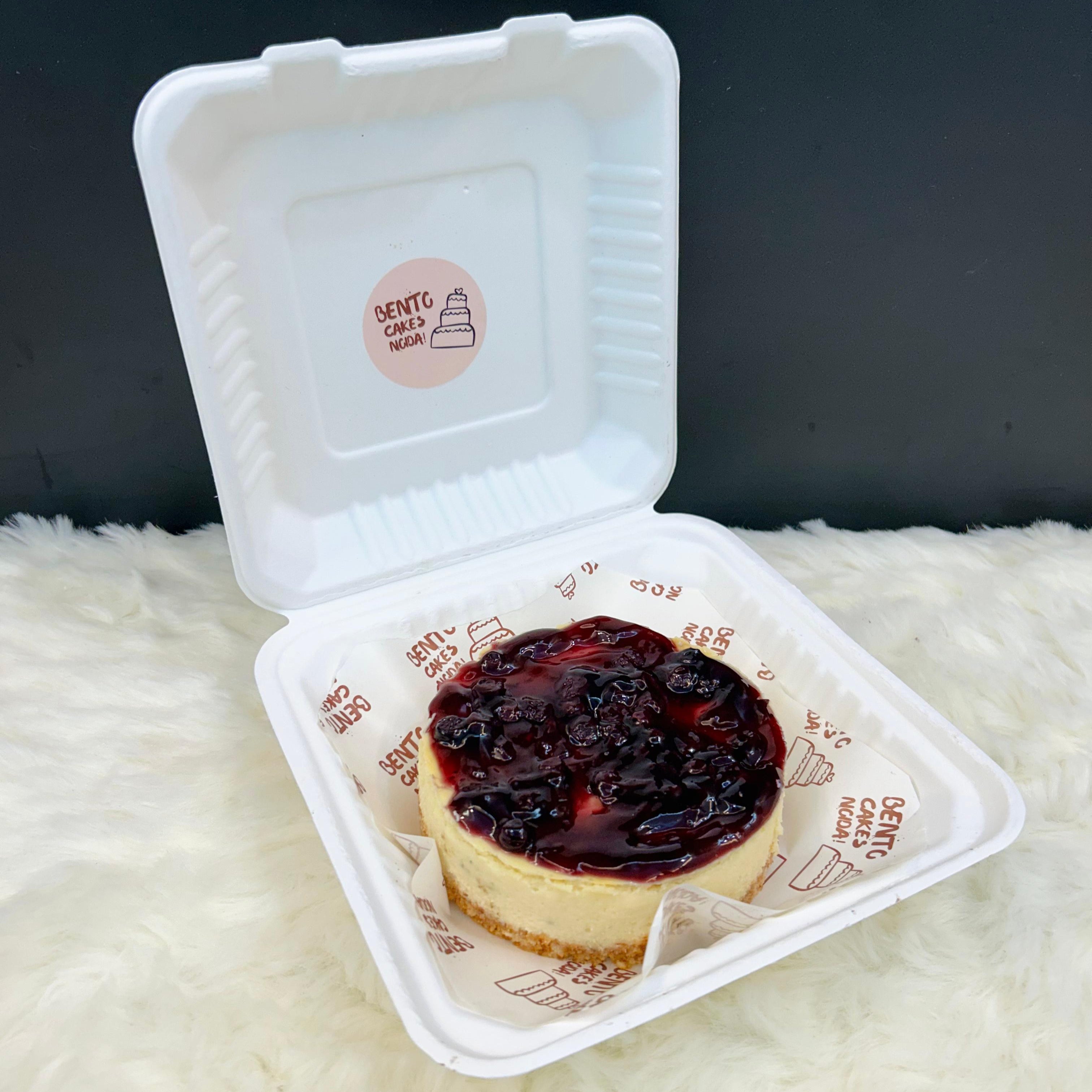 A rich and creamy blueberry cheesecake presented in a white bento-style container, topped with a glossy layer of blueberry compote and garnished with fresh blueberries, set against a minimalistic background.