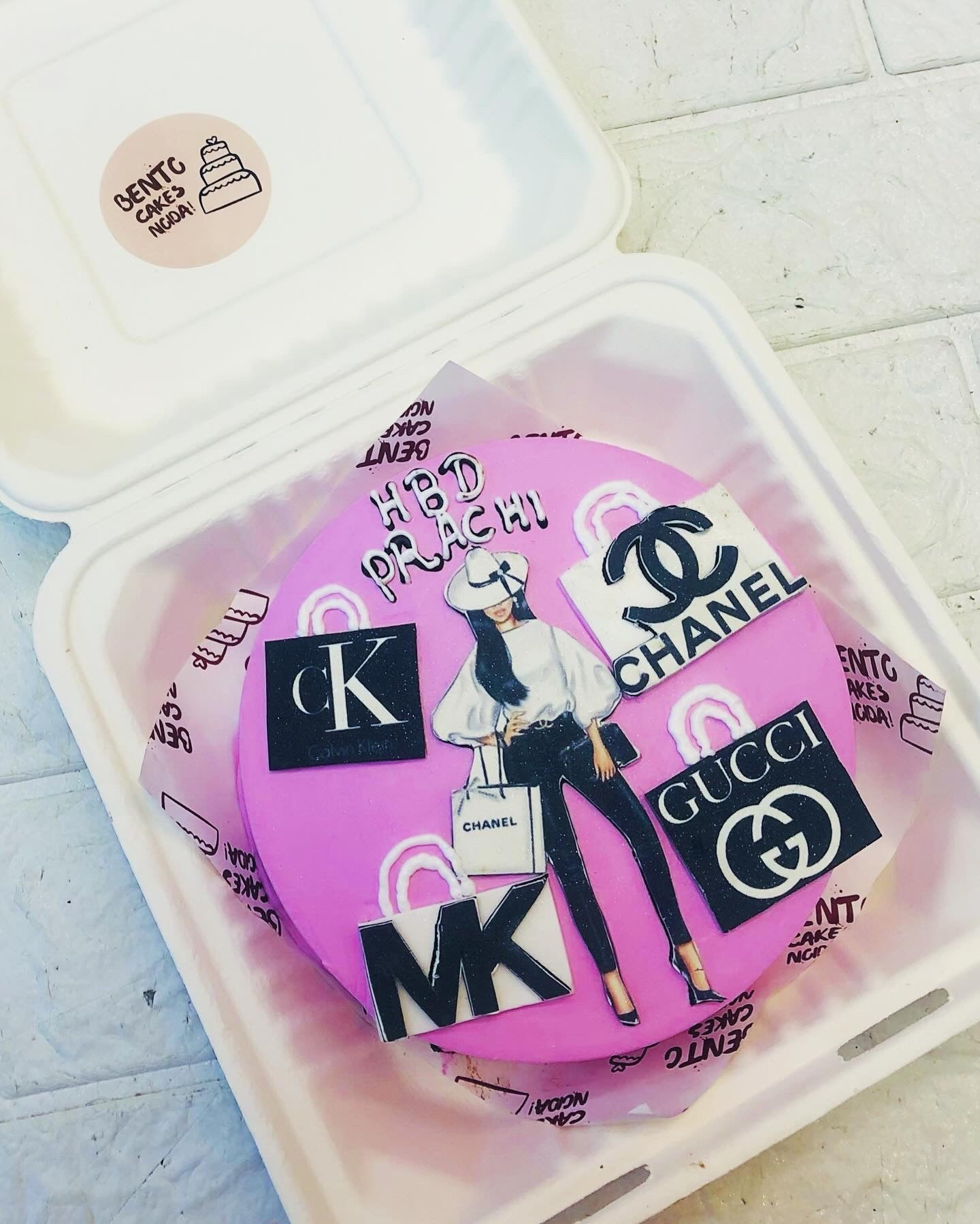 A stylish pink birthday cake featuring a fashion illustration of a woman holding a Chanel shopping bag, surrounded by luxury brand logos like Chanel, Gucci, Calvin Klein, and Michael Kors. The cake has "HBD PRACHI" written on it.