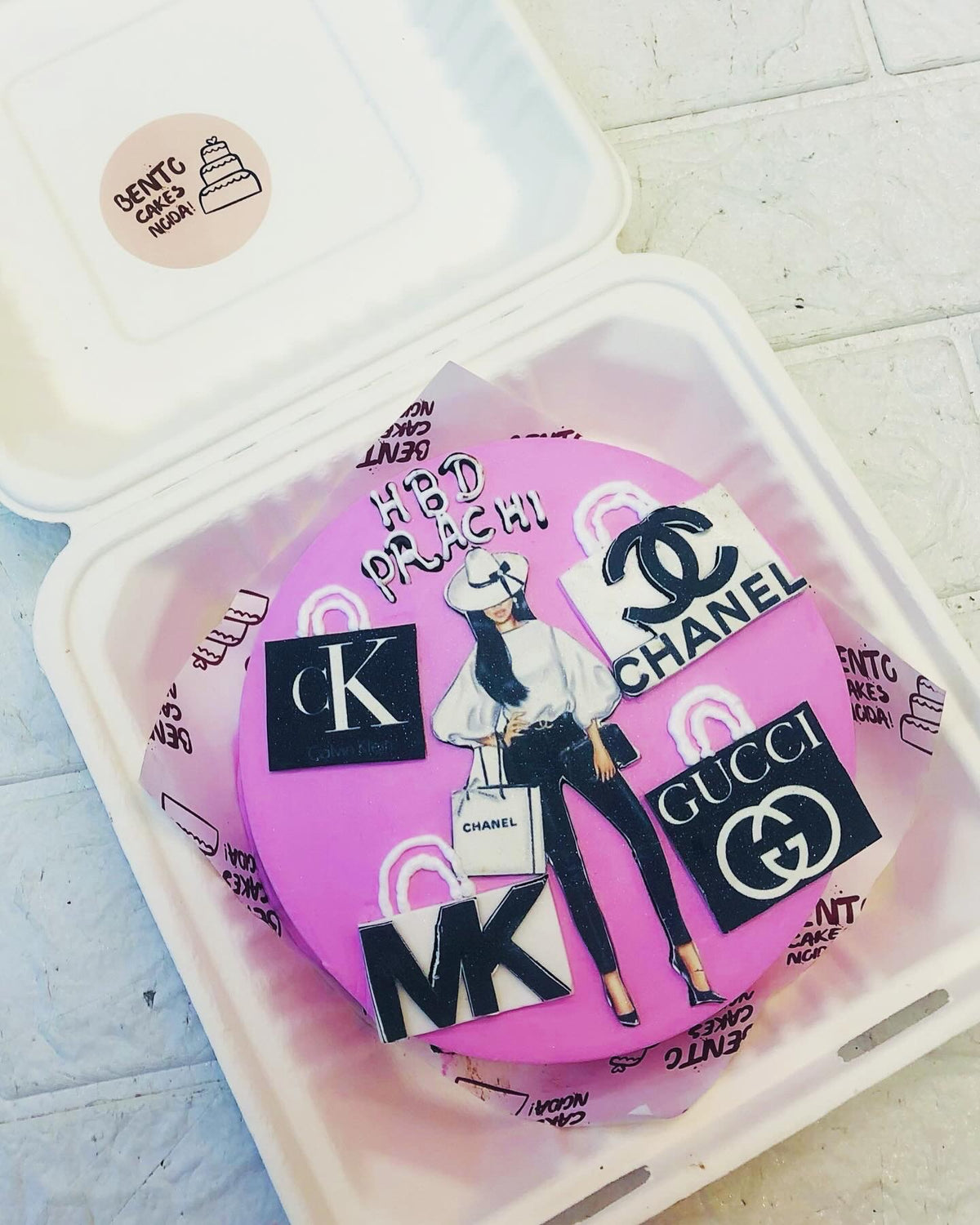A stylish pink birthday cake featuring a fashion illustration of a woman holding a Chanel shopping bag, surrounded by luxury brand logos like Chanel, Gucci, Calvin Klein, and Michael Kors. The cake has "HBD PRACHI" written on it.