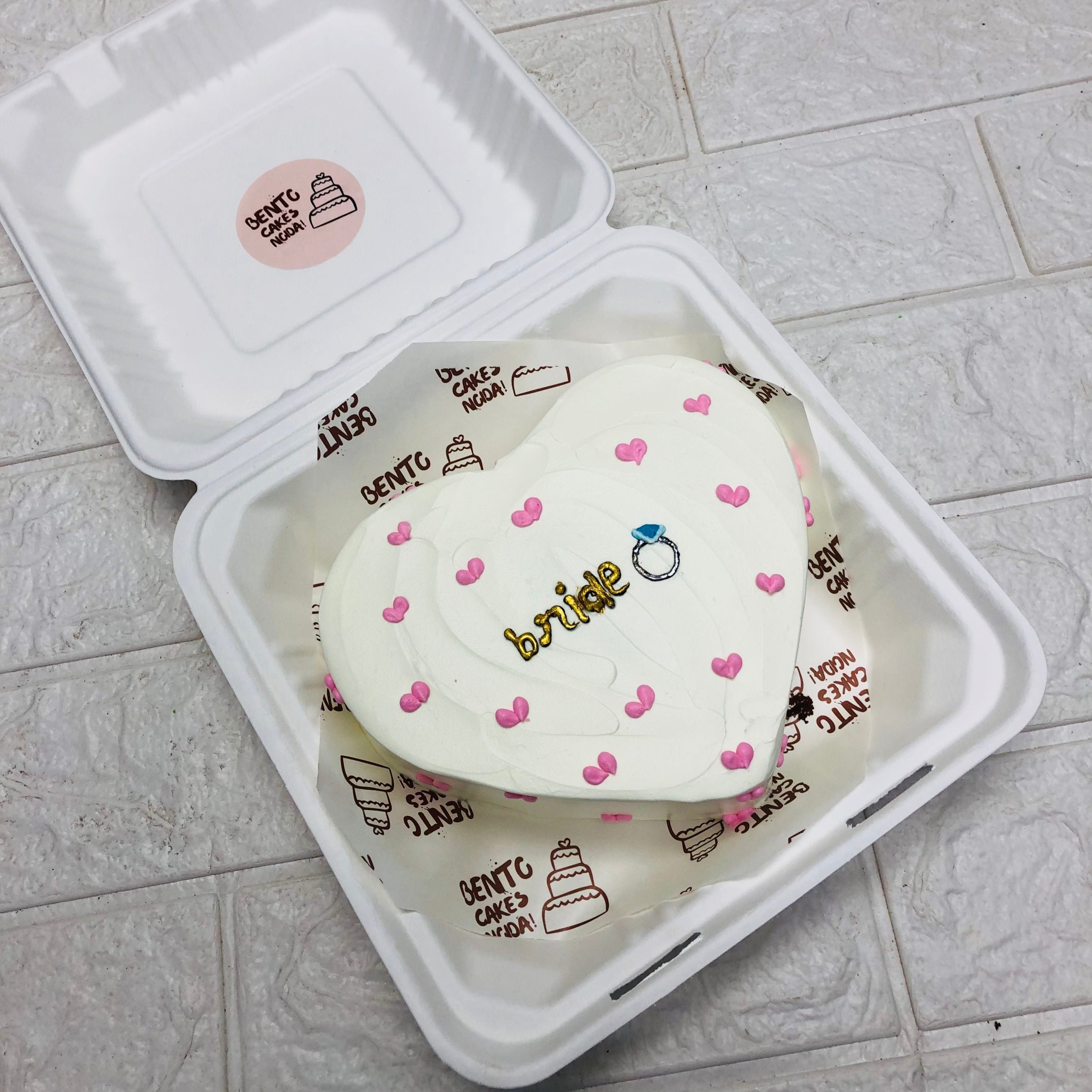 A white heart shape bento cake with cute pink hearts design and a silver ring made on it.