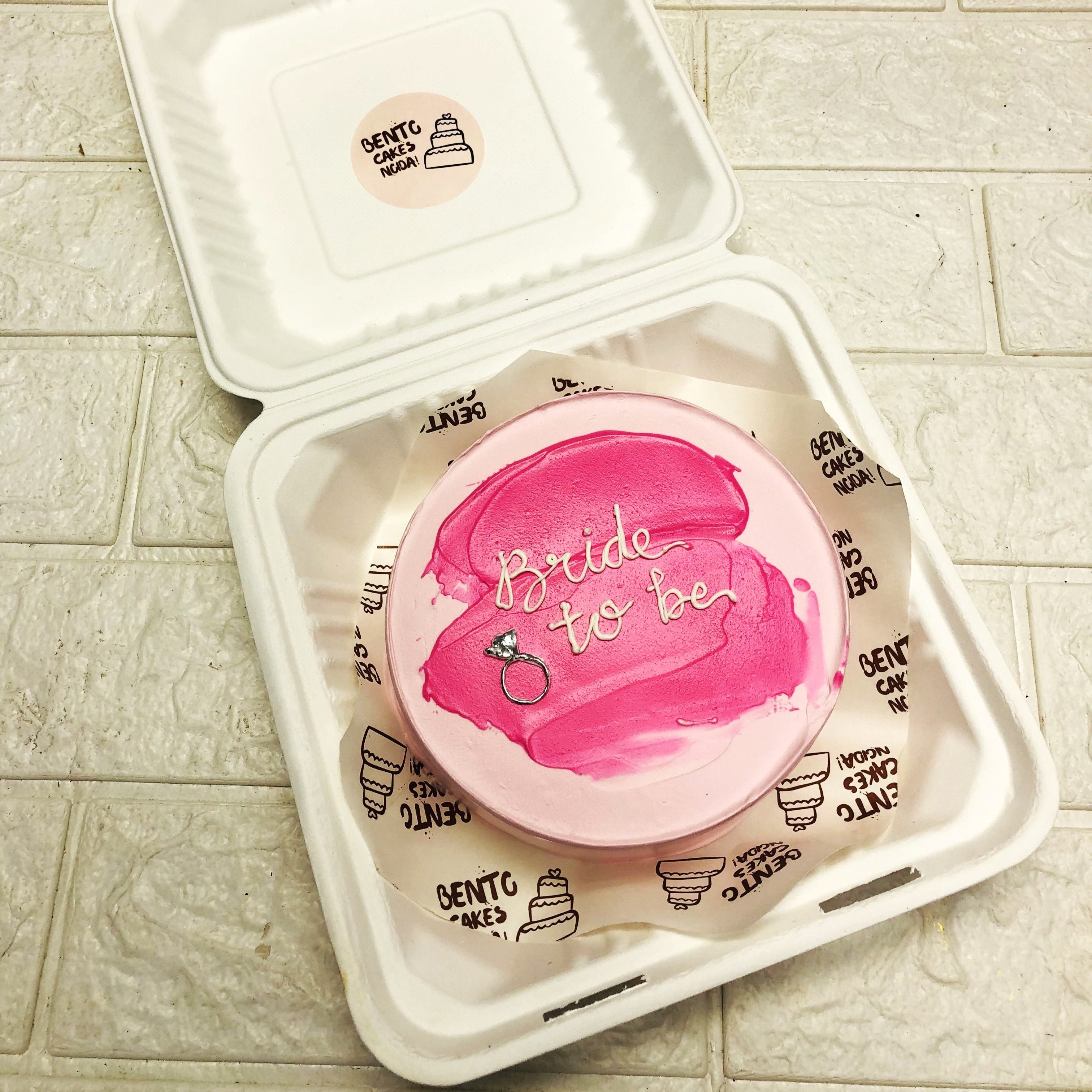 A light pink bento cake having pink shades on top and a silver engagement ring made.