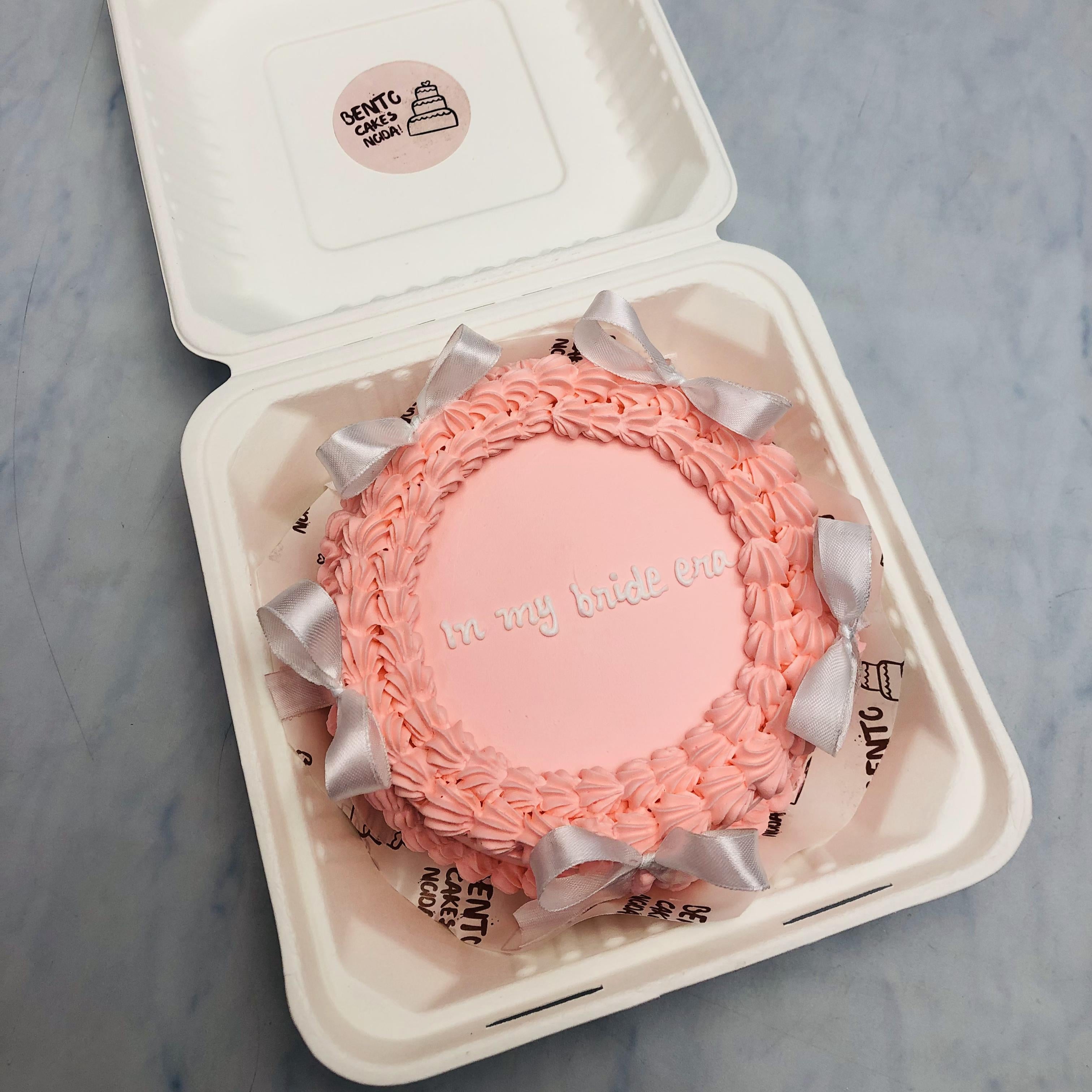 A rose pink round bento cake with white bow designs and a text written with white icing on it.