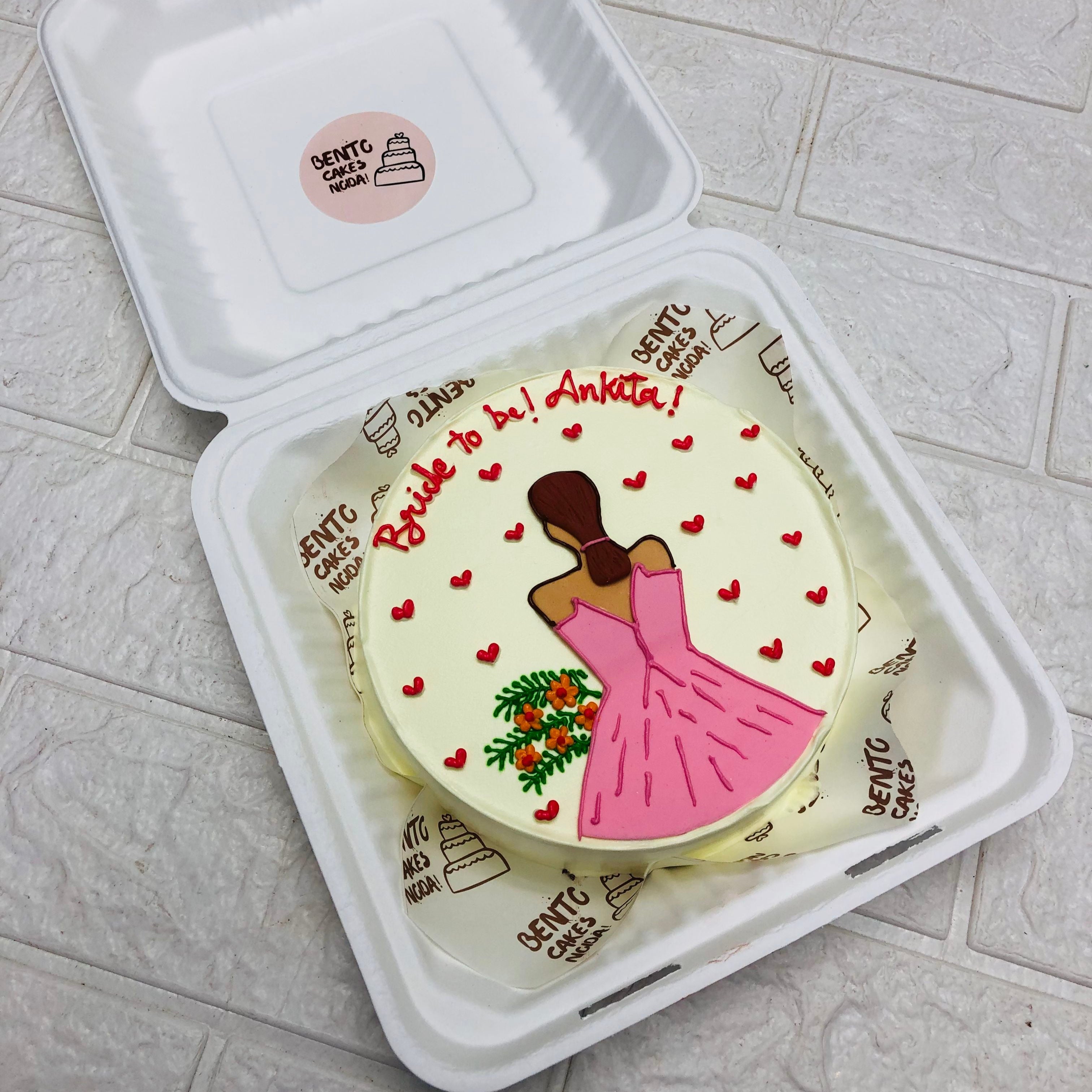 A round white cake with a pink dress design, small red hearts, and the message "Bride to be! Ankita!" written in red icing, placed inside a white takeout box.