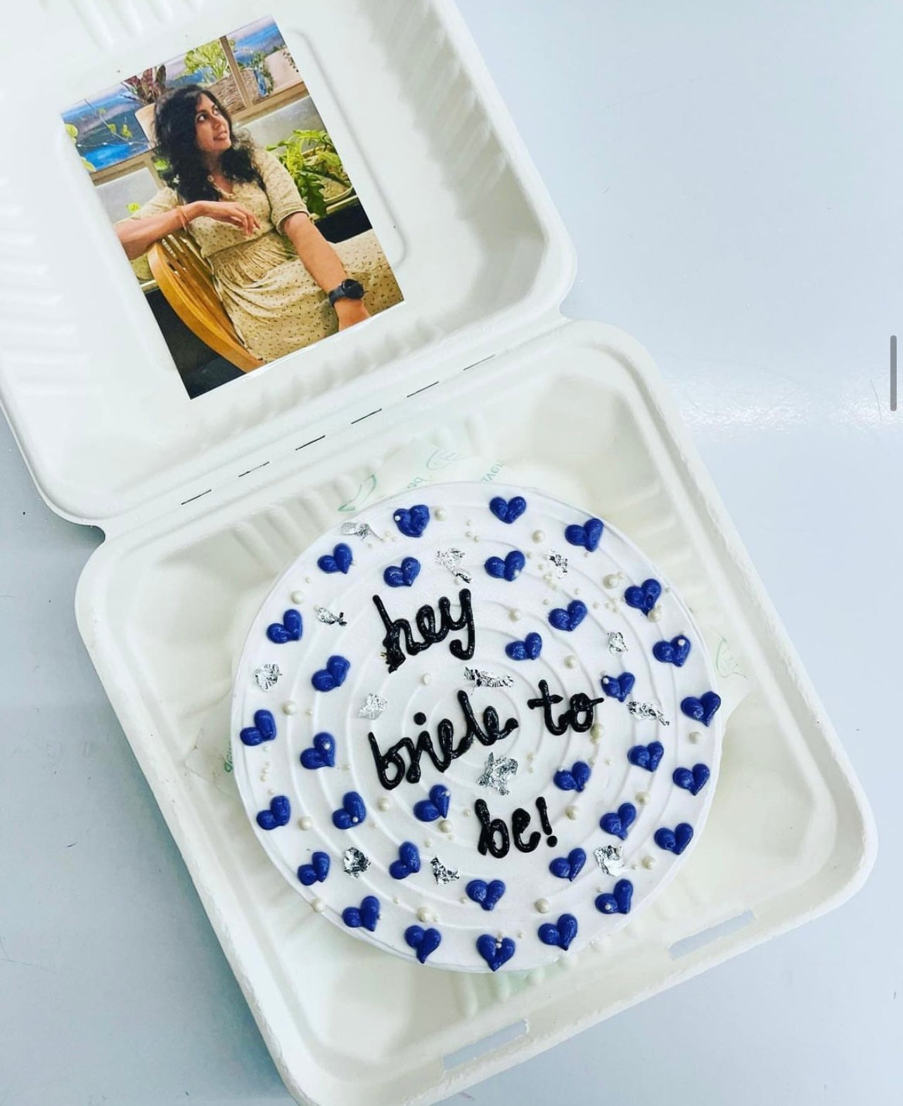 A white cake with blue heart decorations and the text "hey bride to be!" written on it, placed inside a white bento box. The container lid has a photo of a woman sitting on a chair, wearing a light-colored dress.