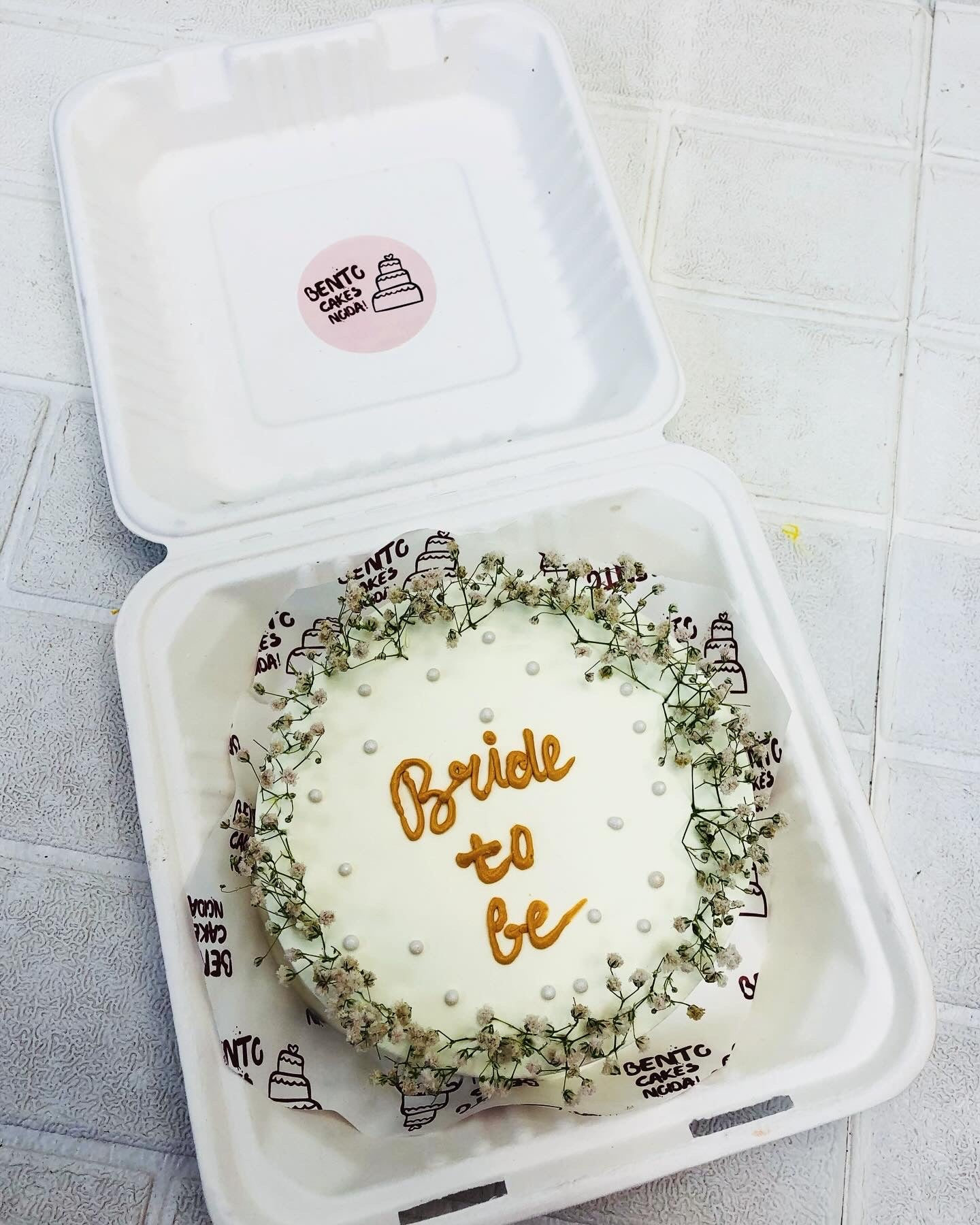 An aesthetic white bento cake with decorative florals and a "Bride to be" ext in center with yellow color.
