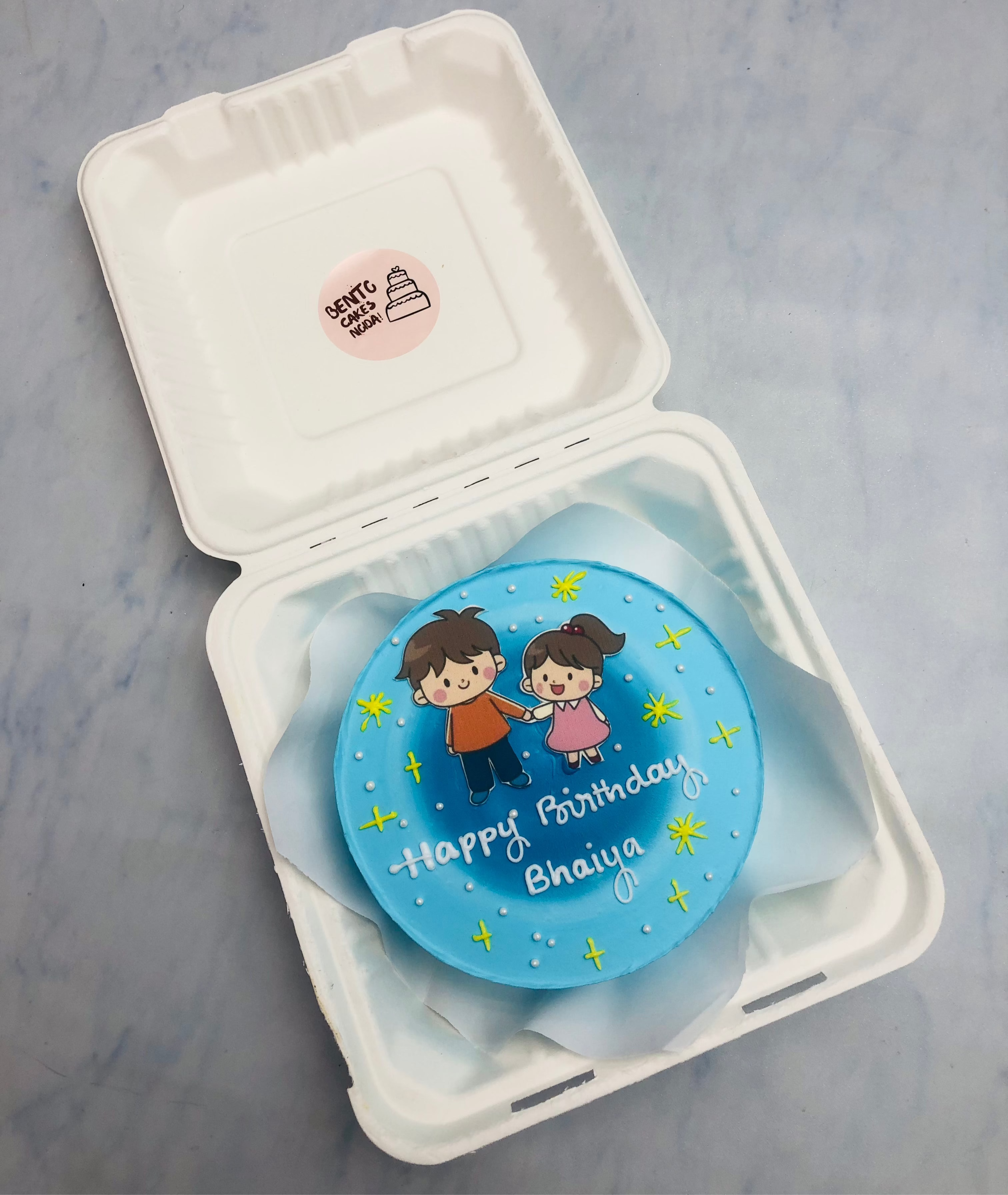 A charming blue bento cake featuring a hand-drawn illustration of a boy and girl holding hands, surrounded by yellow stars, with the message "Happy Birthday Bhaiya" written in white icing.