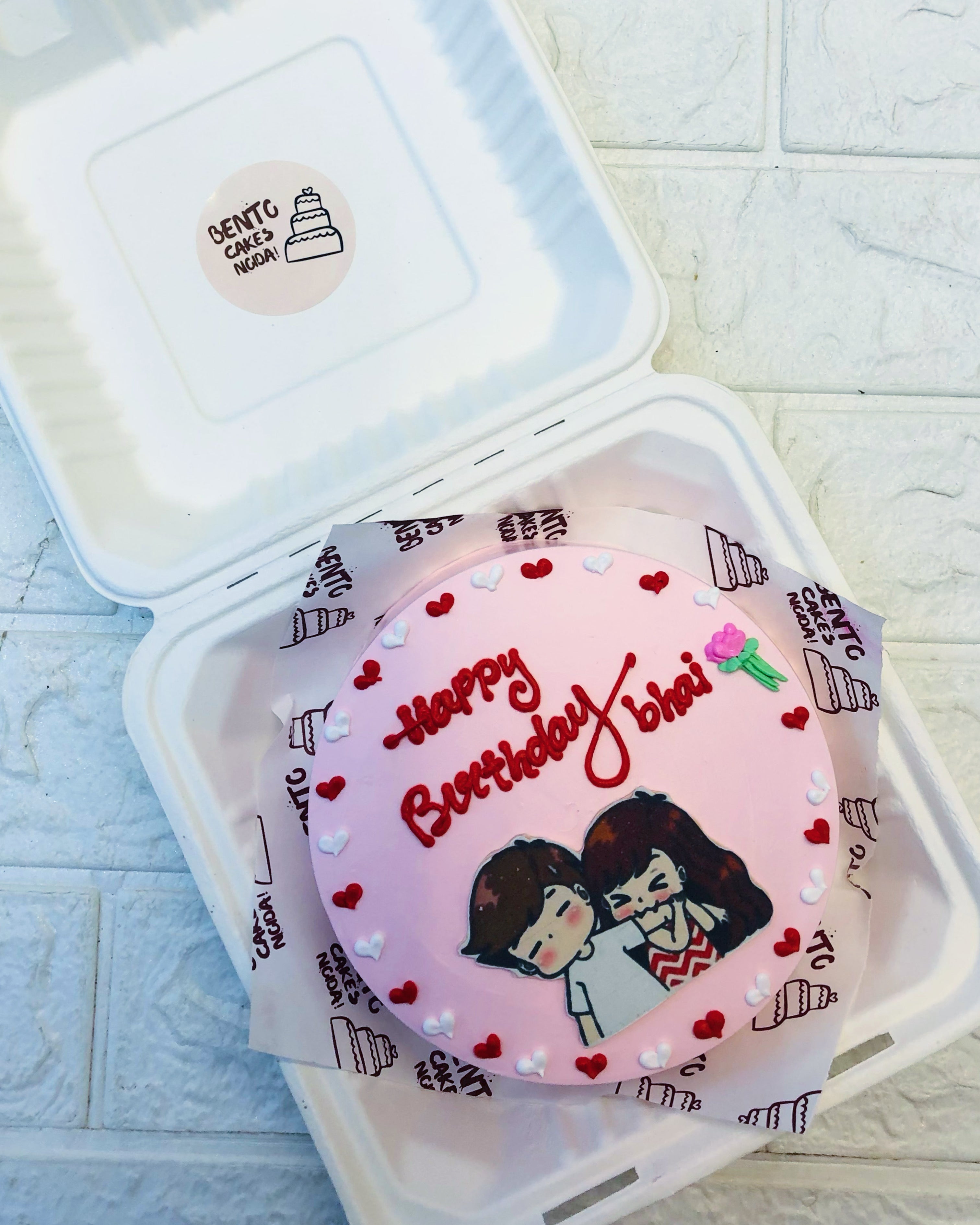A pink bento cake with the message 'Happy Birthday bhai' written in red icing, featuring a cartoon illustration of a boy and girl, surrounded by small red and white heart decorations.