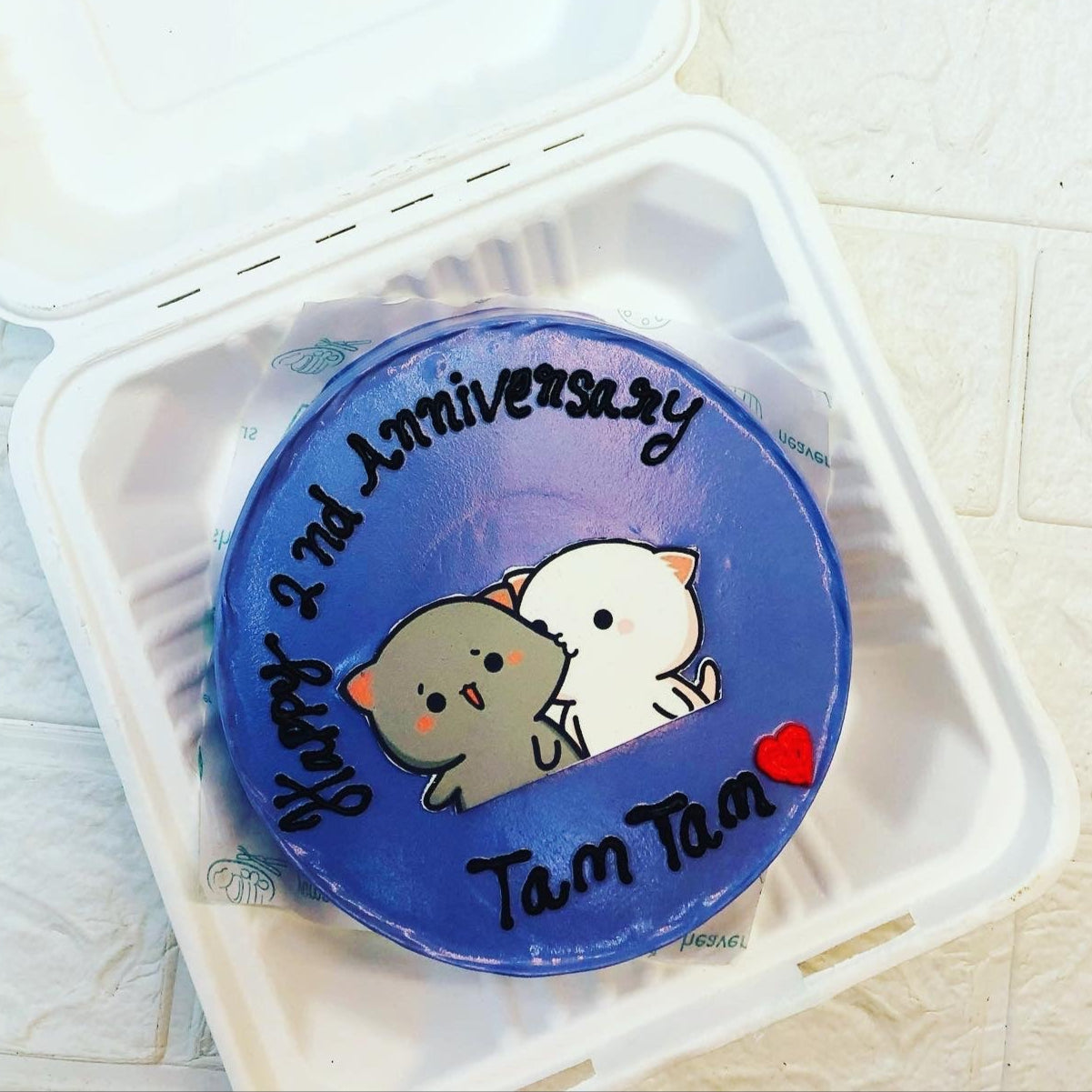 A dark blue icing cake with bubu dudu cartoon sticker in the center and a text "Happy 2nd Anniversary" and "Tam Tam" written on it with black icing and a red color heart also designed on it.