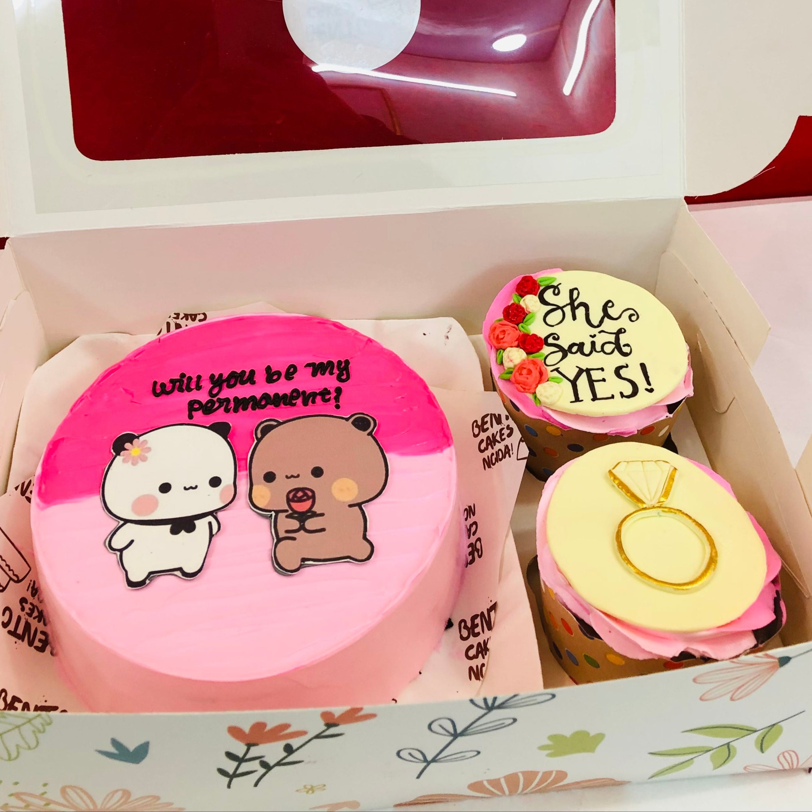 A round pink cake with two cartoon characters, one holding a rose, and the words "Will you be my permanent?" written in elegant black icing. Accompanied by two cupcakes, one featuring "She said YES!" and another with a ring design.