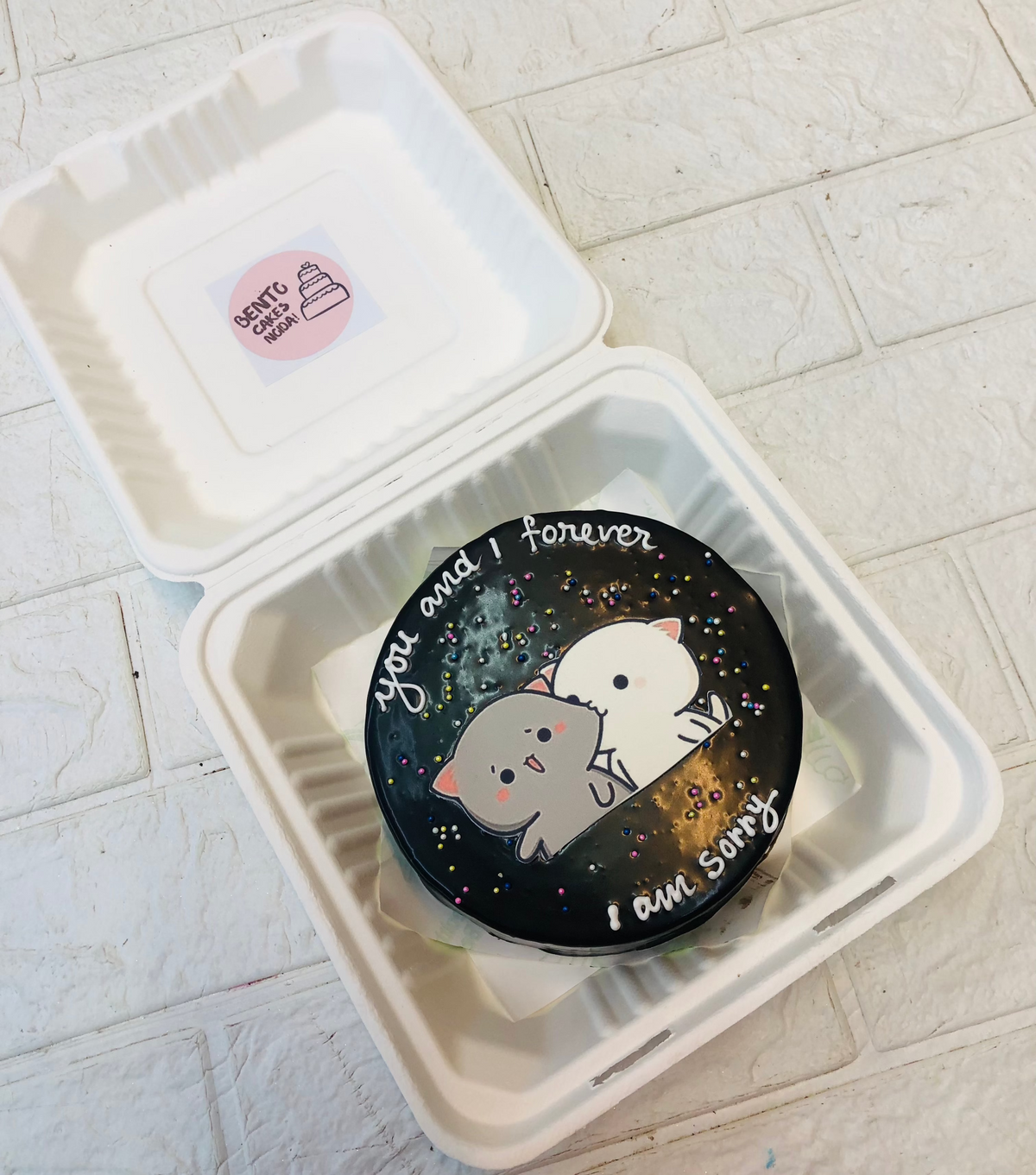 A chocolaty black bento cake having a kissing bubu dudu print and decorated with colorful sprinkles.