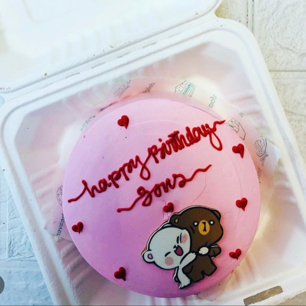 A pink birthday cake with red icing that reads "happy birthday Jons" and is decorated with small red hearts and a cute illustration of two hugging bears, one white and one brown. The cake is placed inside a white takeout container.