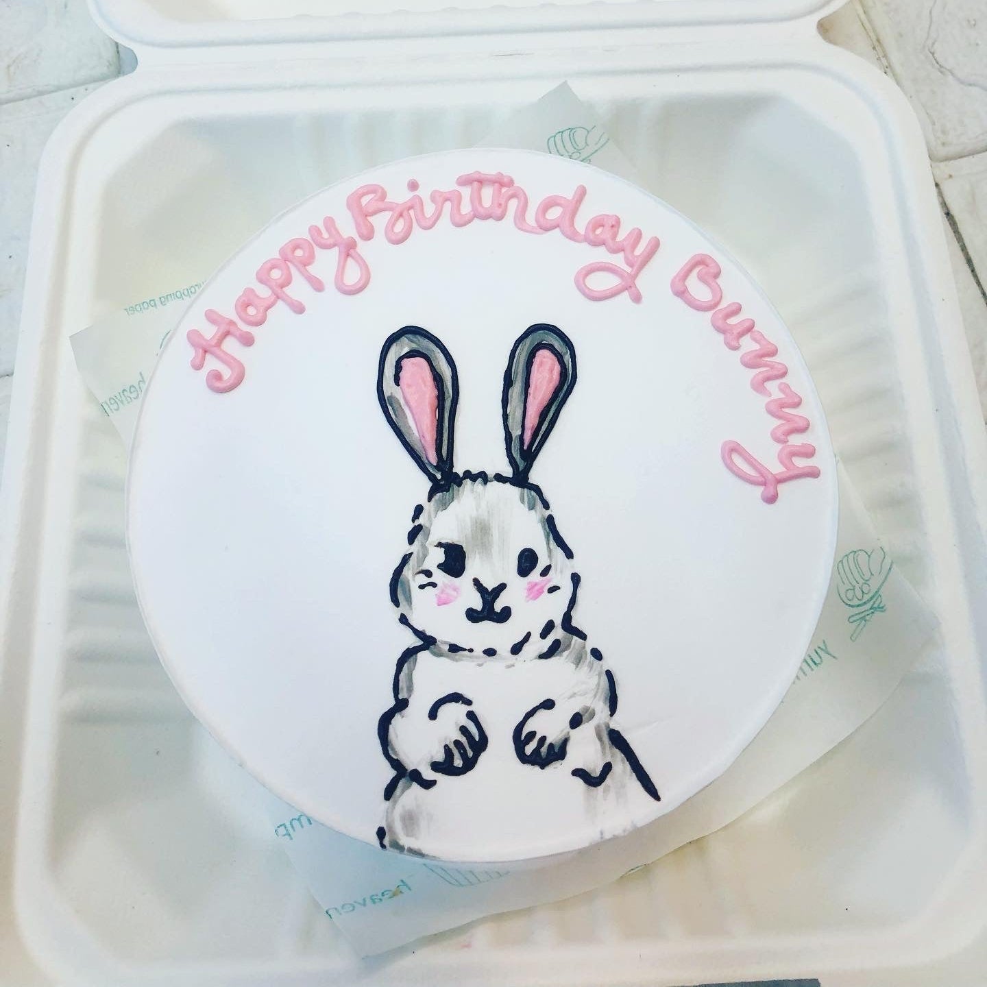 A colorful birthday cake topped with a cute bunny, all neatly placed in a clear plastic container.
