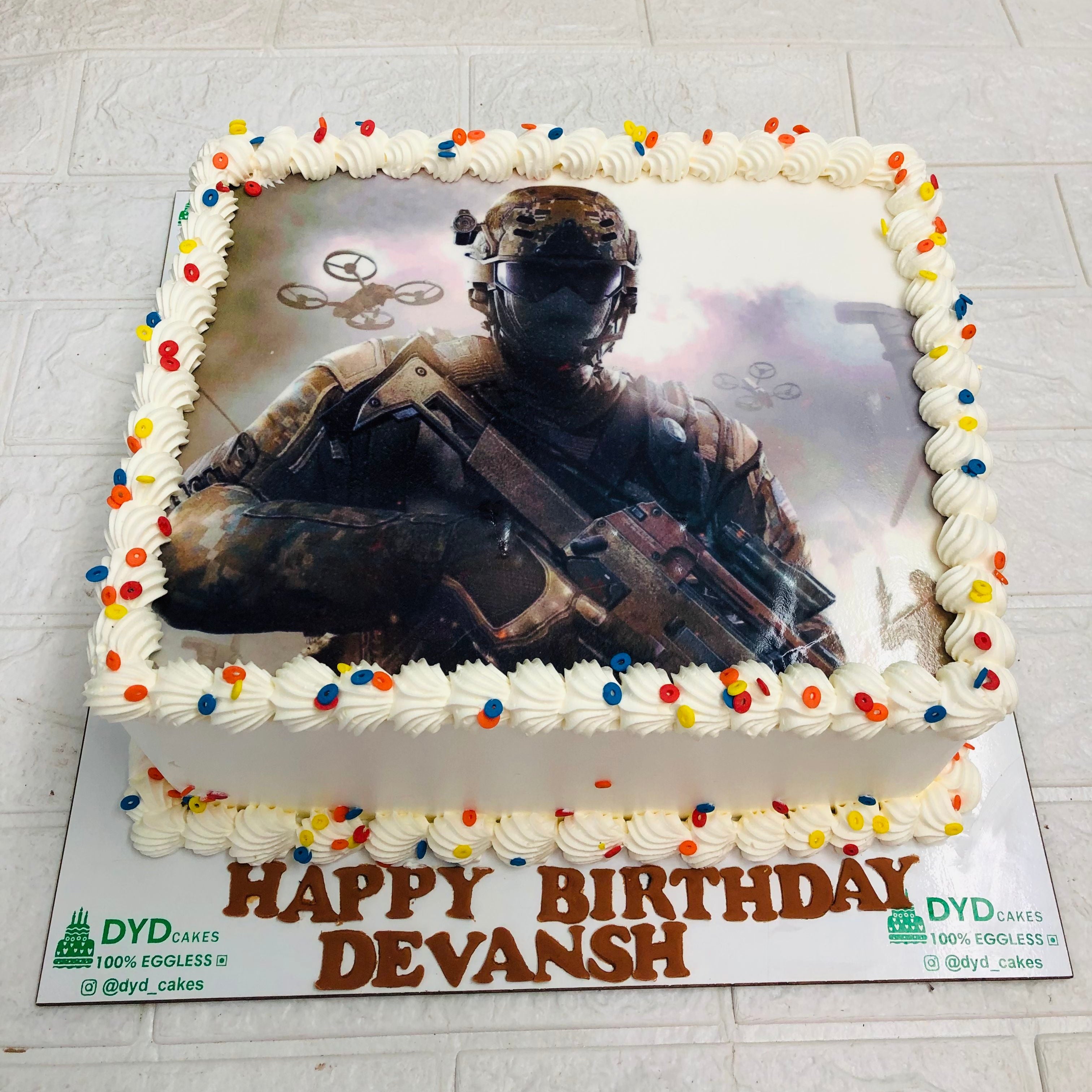 A rectangular birthday cake with white frosting and colorful sprinkles, featuring an edible image of a soldier in combat gear with drones in the background. The text "HAPPY BIRTHDAY DEVANSH" is written at the base in brown icing.