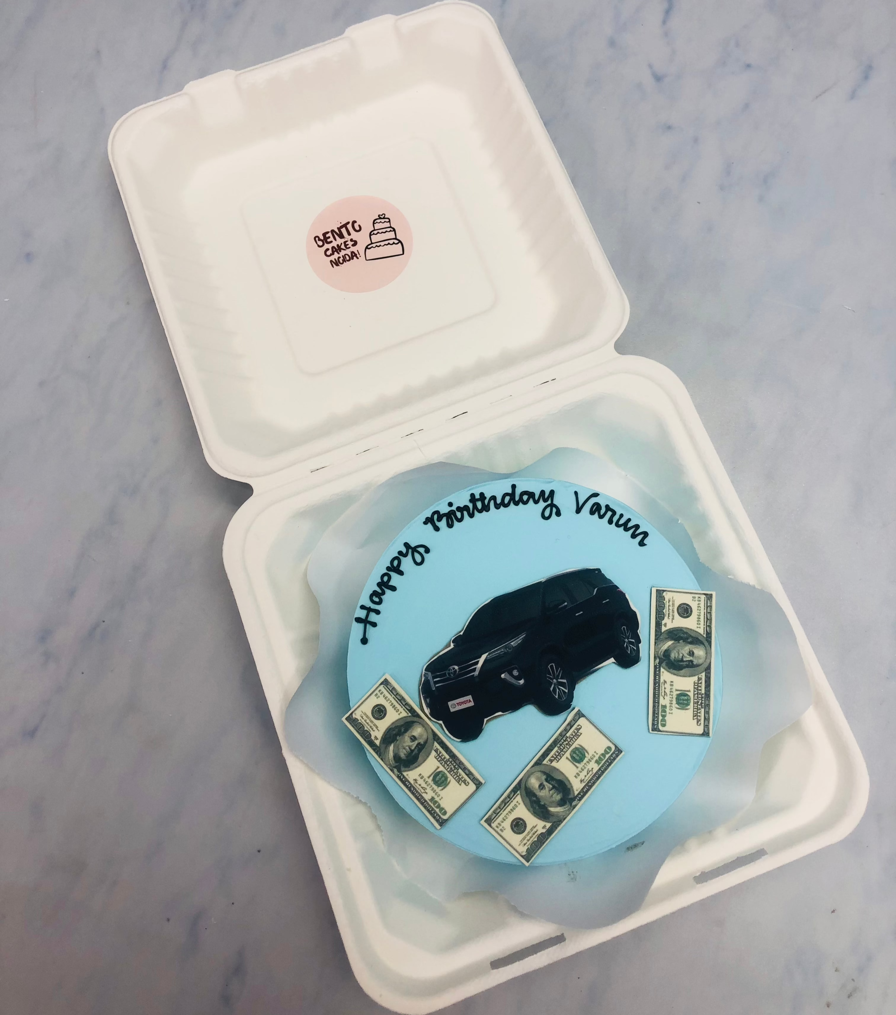 A light blue bento cake with Black Fortuner car and Dollor photo prints.