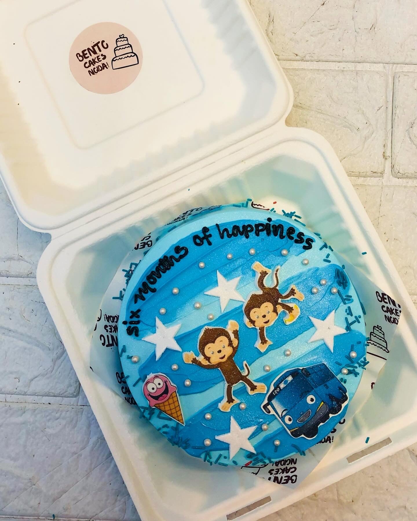 A light blue cake designed with blue hades on top with cute monkeys, bus and icecream stickers on it.