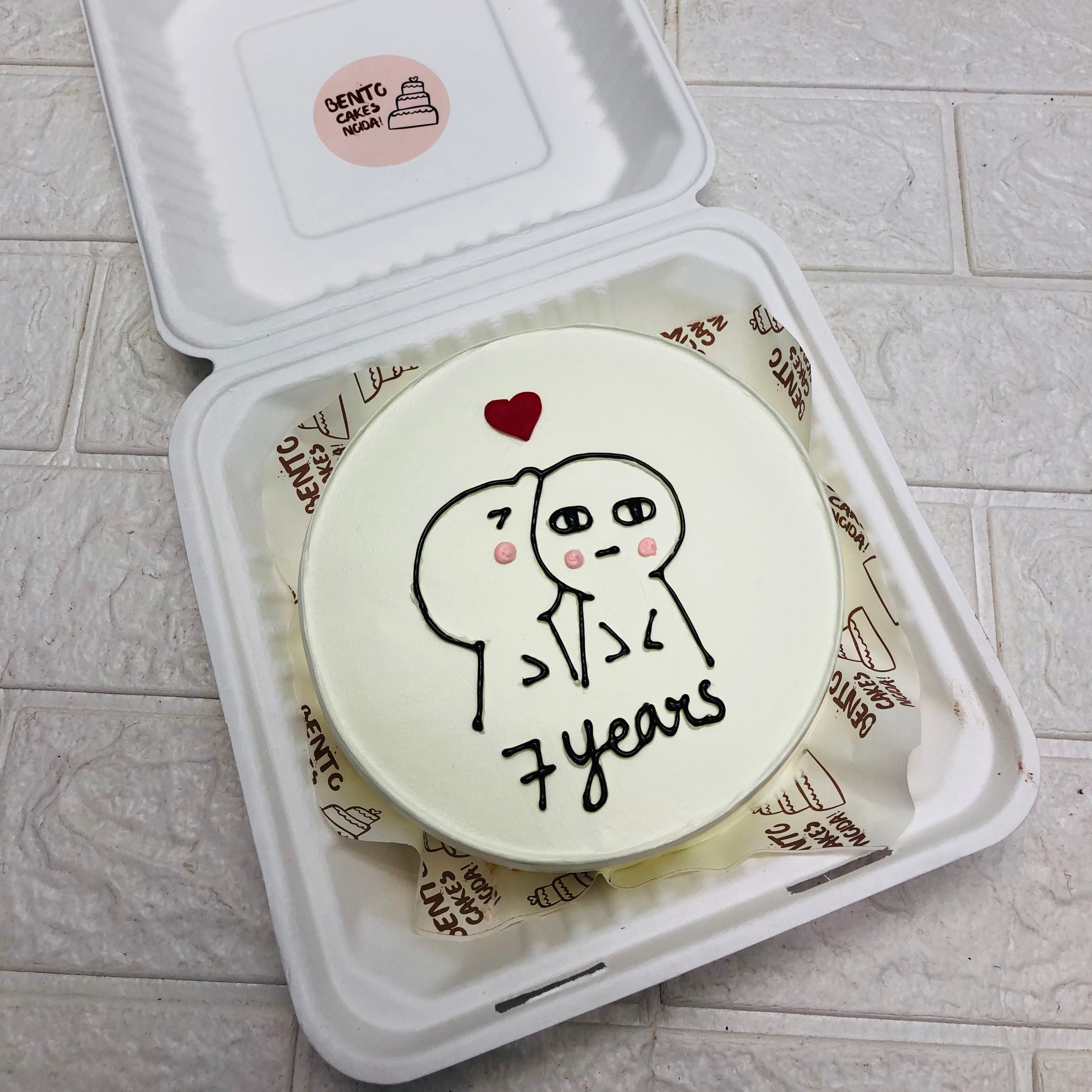 A white cake designed with 2 cartoon characters kissing and a red heart design created on their head and a text "7 Year" written with black icing below cartoon. This cake is designed for marriage anniversary occasions.