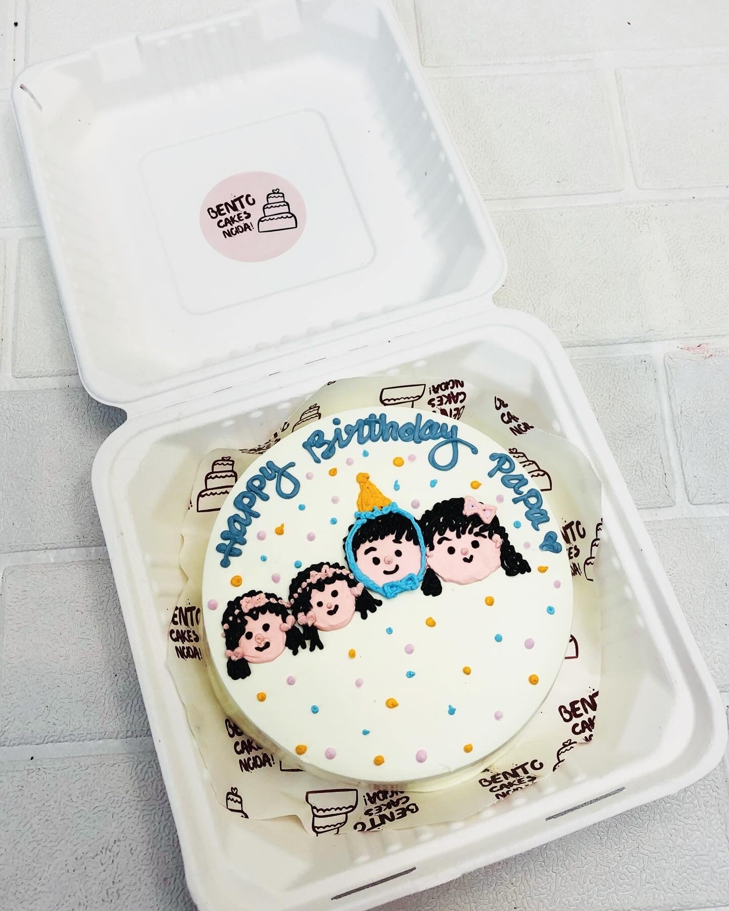 A white bento cake with blue decorations, featuring a simple drawing of a person's face wearing a party hat, surrounded by small blue hearts. The cake is inscribed with the text 'HAPPY BIRTHDAY PAPA' in blue icing. The cake is placed inside a white bento box.